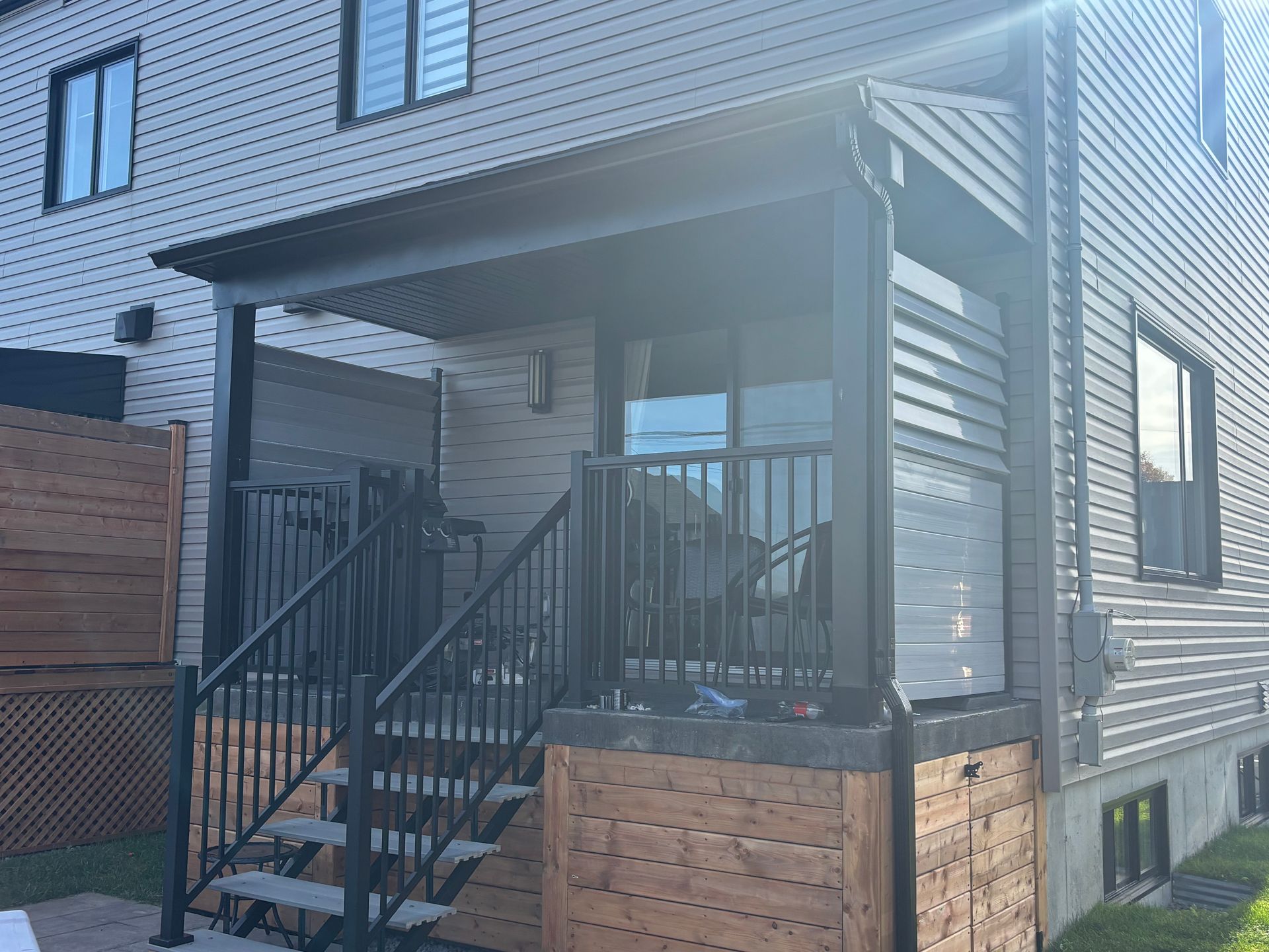 Terrasse arrière avec escalier, rambarde noire, éléments en bois et porche couvert.