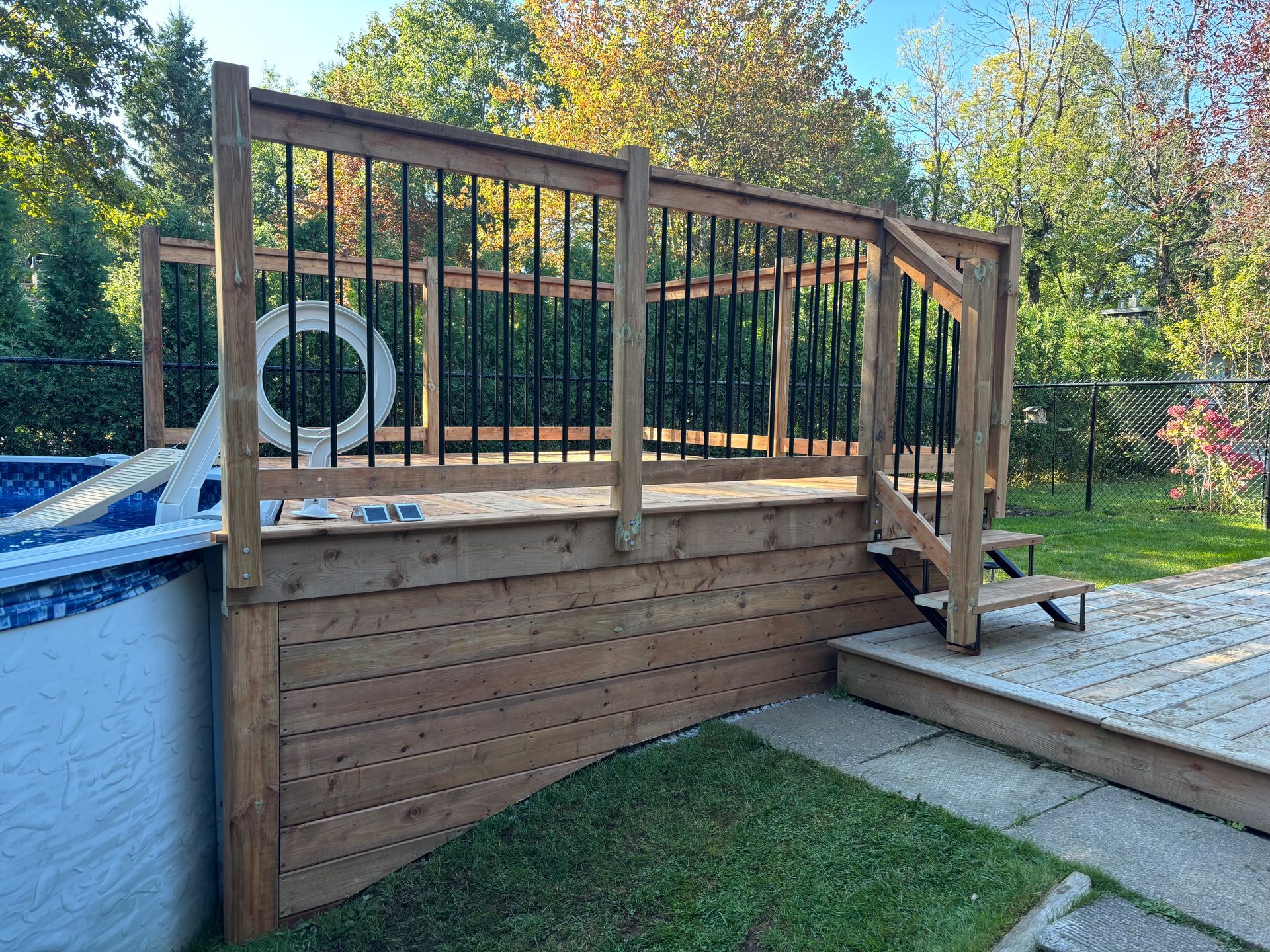 Terrasse en bois avec balustrades noires entourant une piscine hors sol ; des marches mènent à une terrasse inférieure.