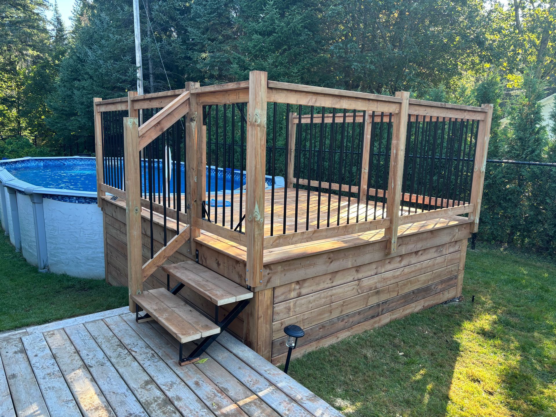 Terrasse en bois avec marches, rambarde noire et piscine en arrière-plan.