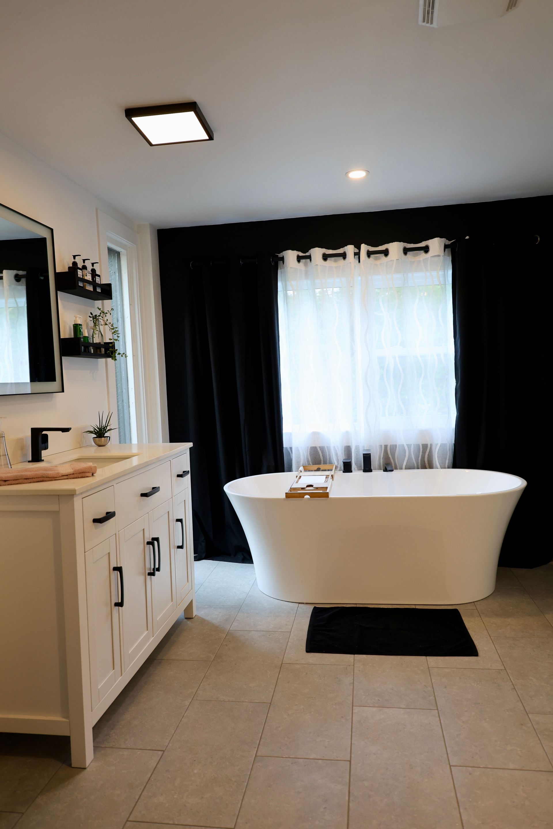 Salle de bains avec baignoire îlot blanche, meuble-lavabo, rideaux noirs et carrelage gris clair au sol.