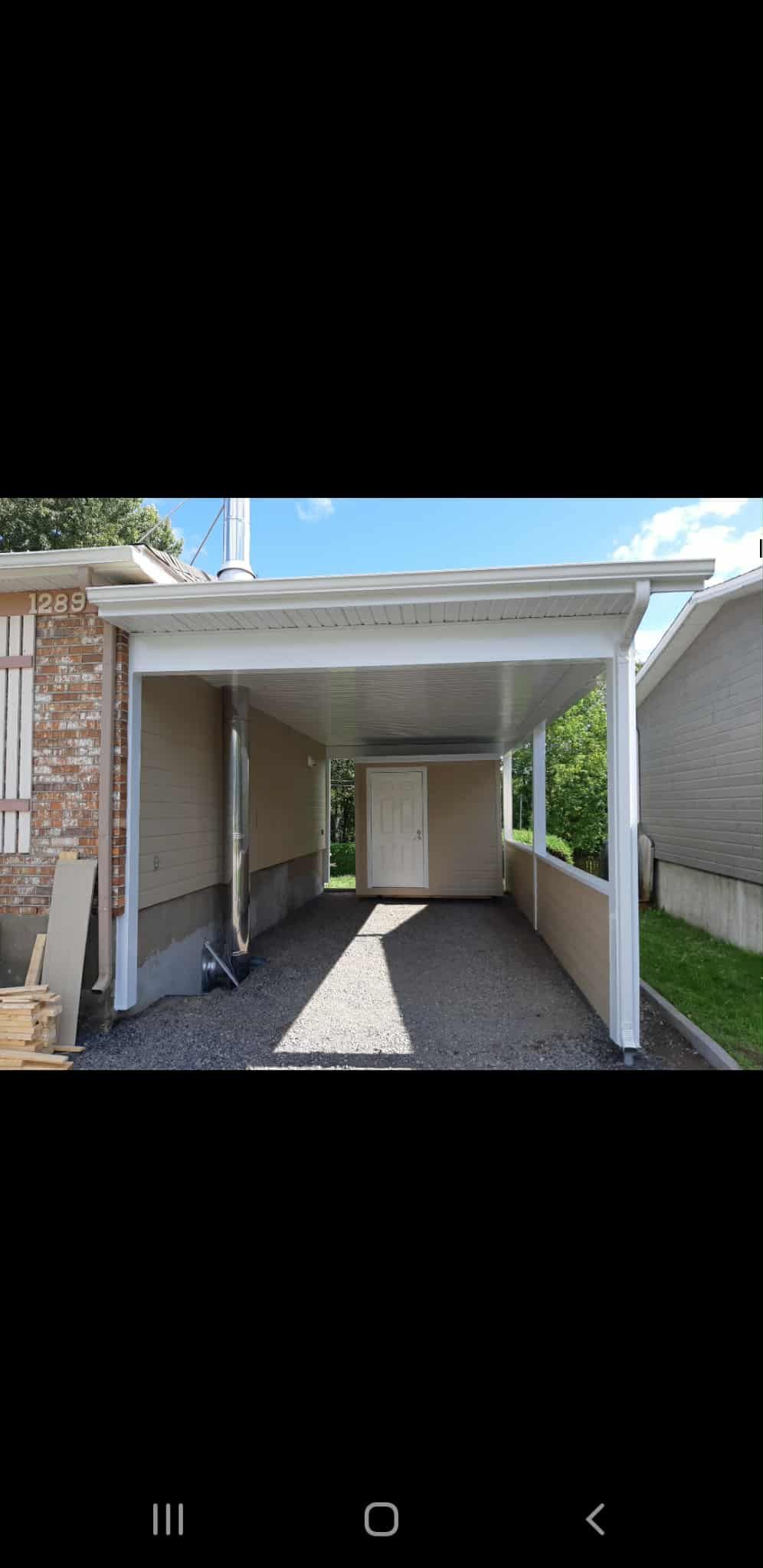 Un abri de voiture attenant à un bâtiment en briques. Allée en gravier, murs beiges et structure blanche.