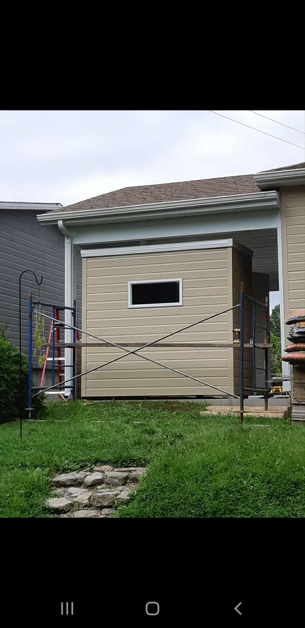 Un abri beige avec une fenêtre, dans un jardin herbeux. Un chemin de pierre y mène. Ciel nuageux au-dessus.