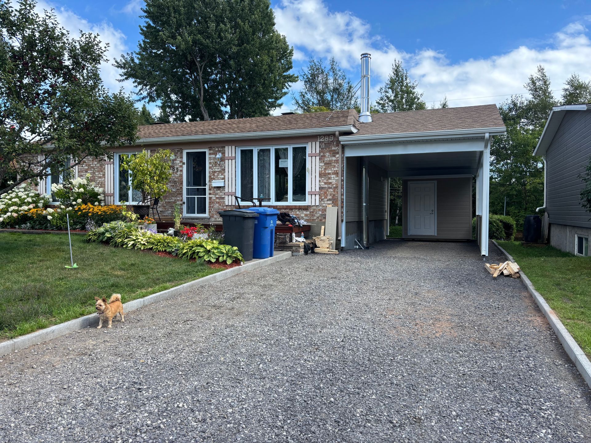 Maison de plain-pied de style ranch avec allée gravillonnée, abri voiture et jardin paysager à l'avant. Un petit chien se tient sur l'allée.