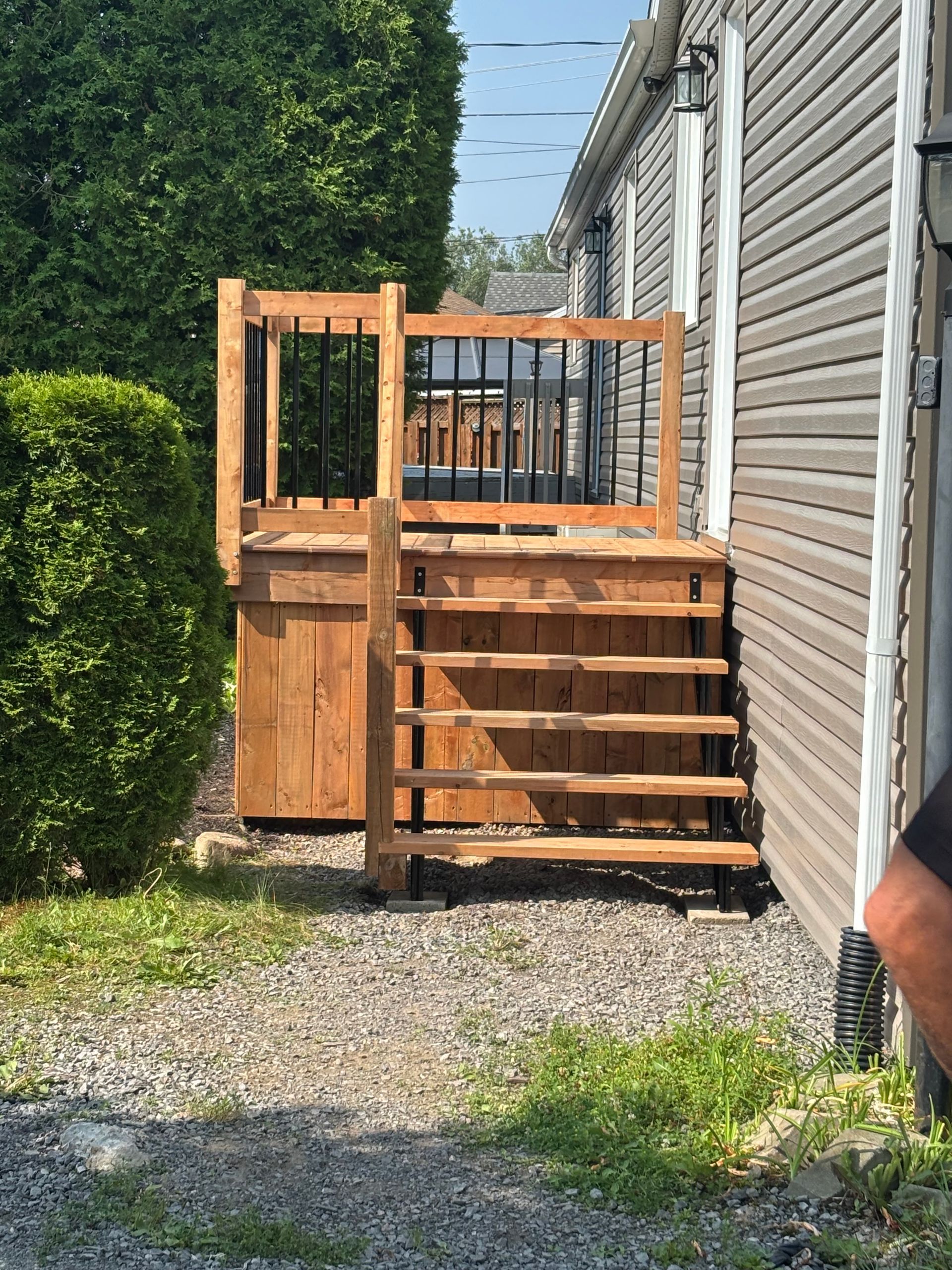 Un escalier extérieur en bois mène à une petite terrasse avec une rambarde noire adossée à un bâtiment.
