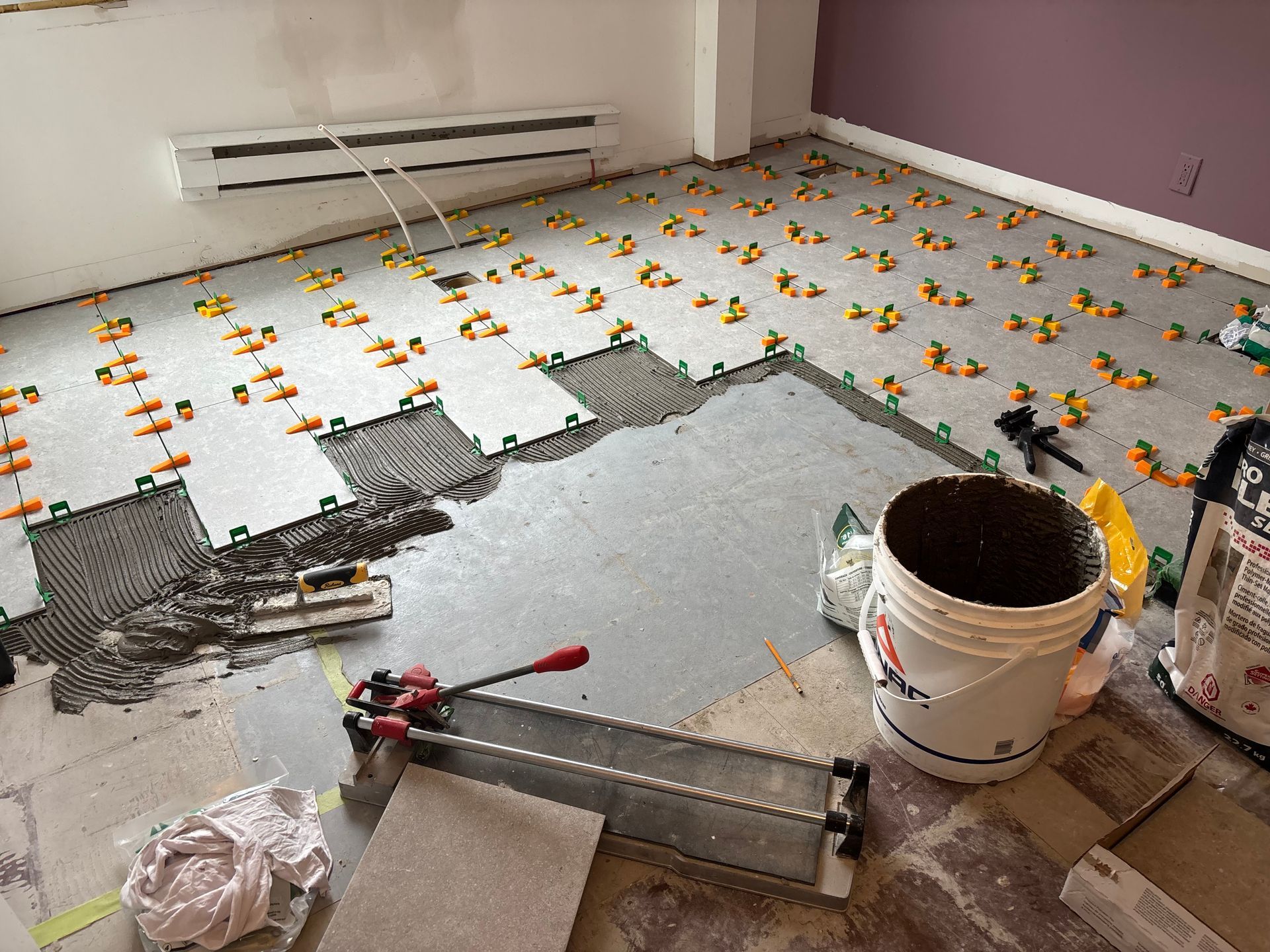 Pièce en cours de pose de carrelage ; sol recouvert de carreaux, croisillons, outils et un seau.