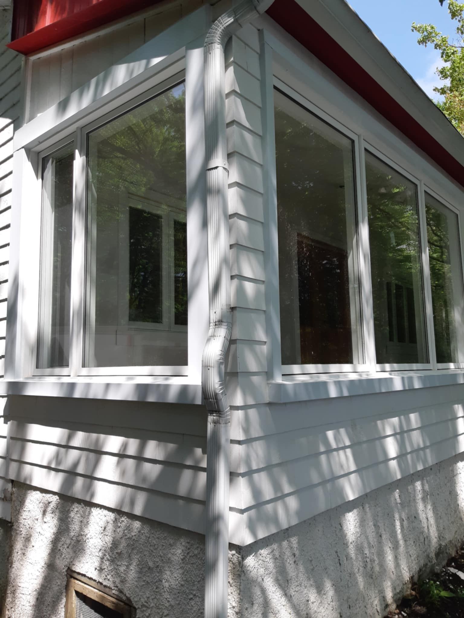 Maison blanche d'angle avec de grandes fenêtres, un bardage blanc, des finitions rouges et une descente de gouttière.