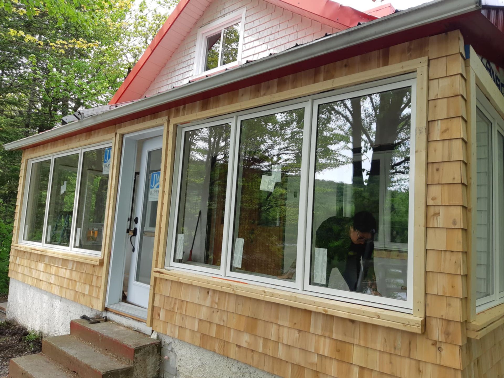 Maison au toit de bardeaux de bois, grandes fenêtres, et une personne qui travaille à l'intérieur. Toit rouge, marches grises.