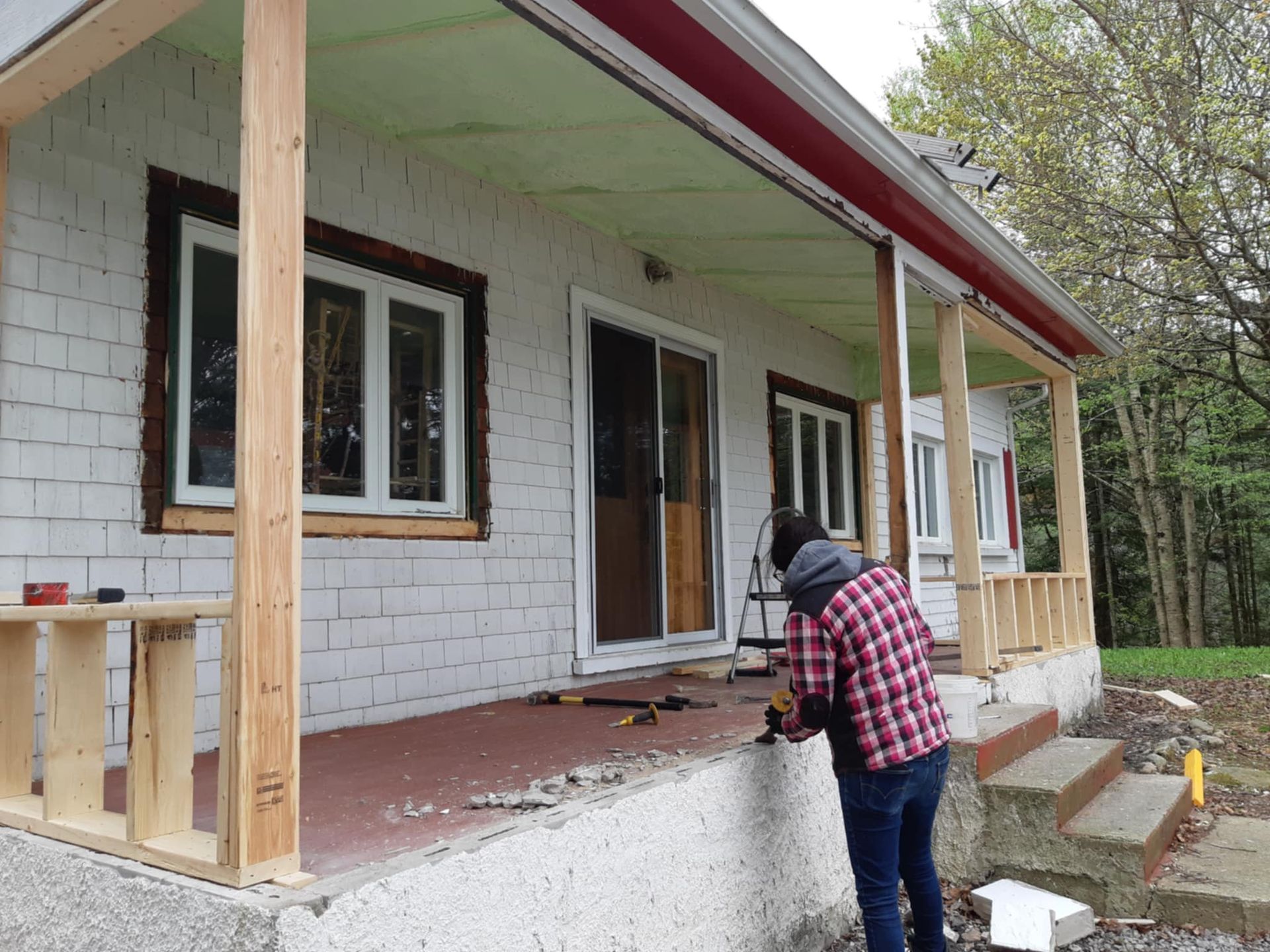 Une personne travaille sur le porche d'une maison en briques blanches, dont la rambarde est neuve et en bois.