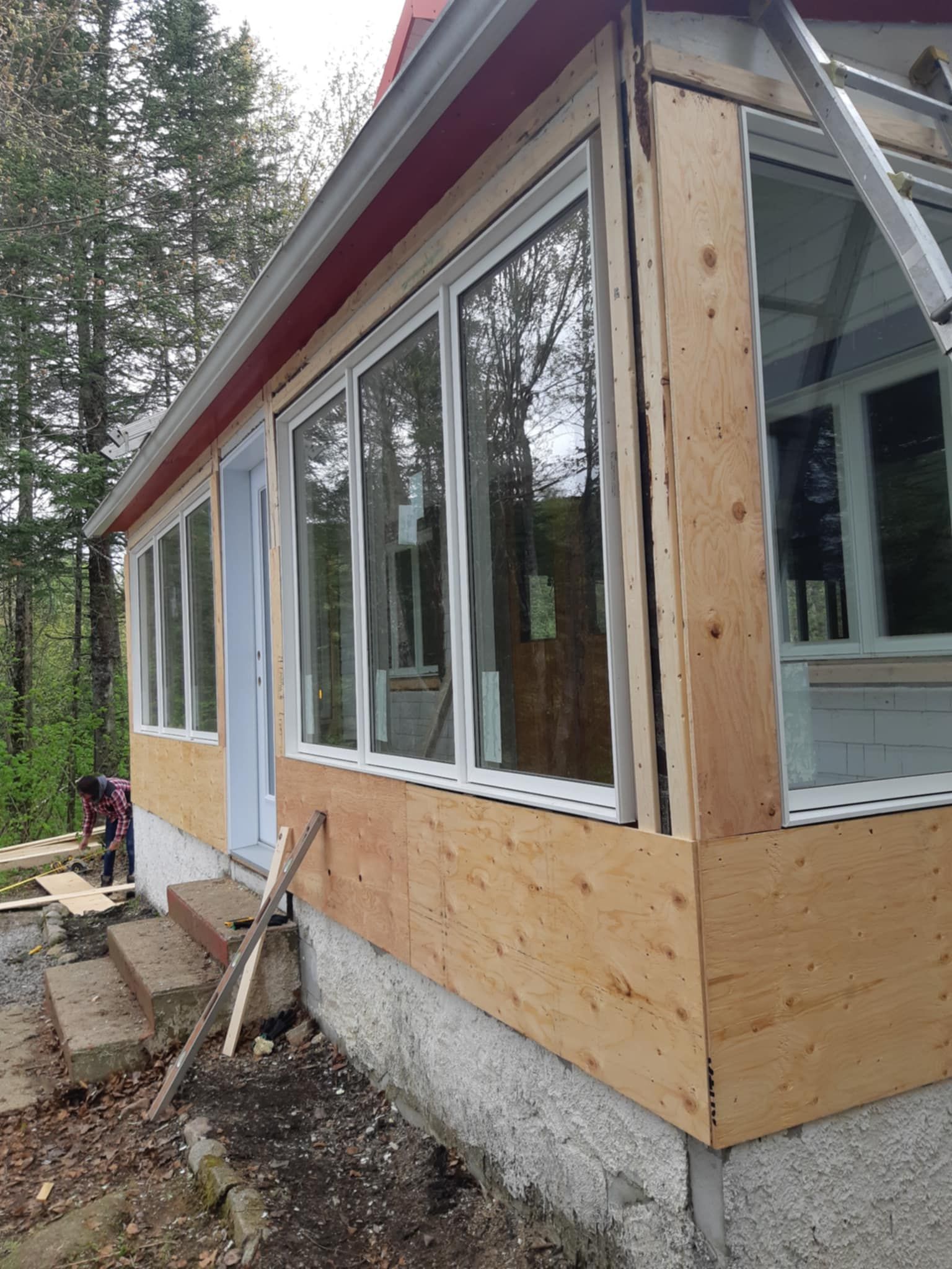 Extérieur d'une maison en rénovation. Bardage en contreplaqué, grandes fenêtres, boiseries rouges. Échelle visible.