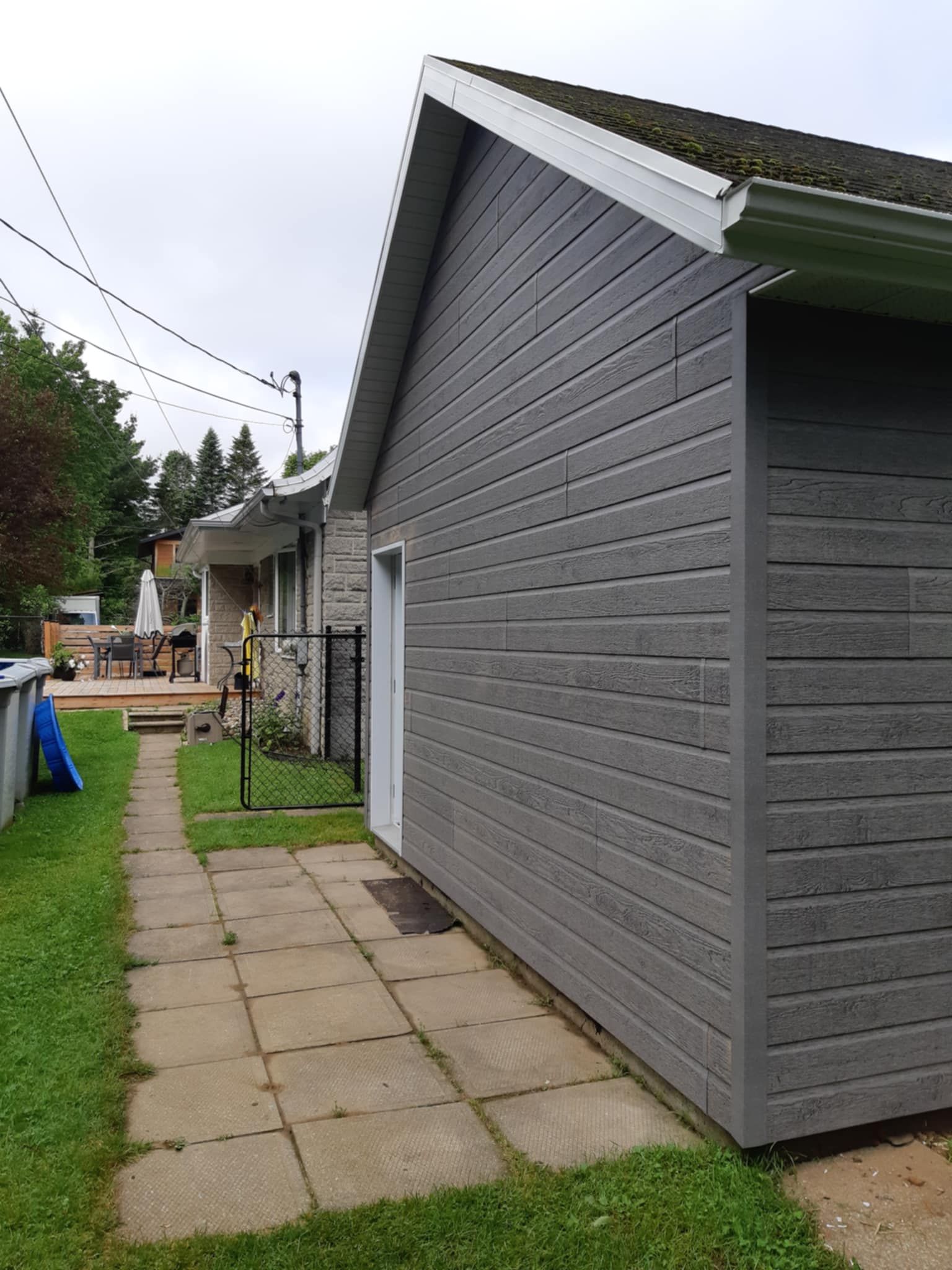 Bâtiment aux murs gris et à la porte blanche, jouxtant un bâtiment en pierre avec un chemin et une terrasse à l'arrière.