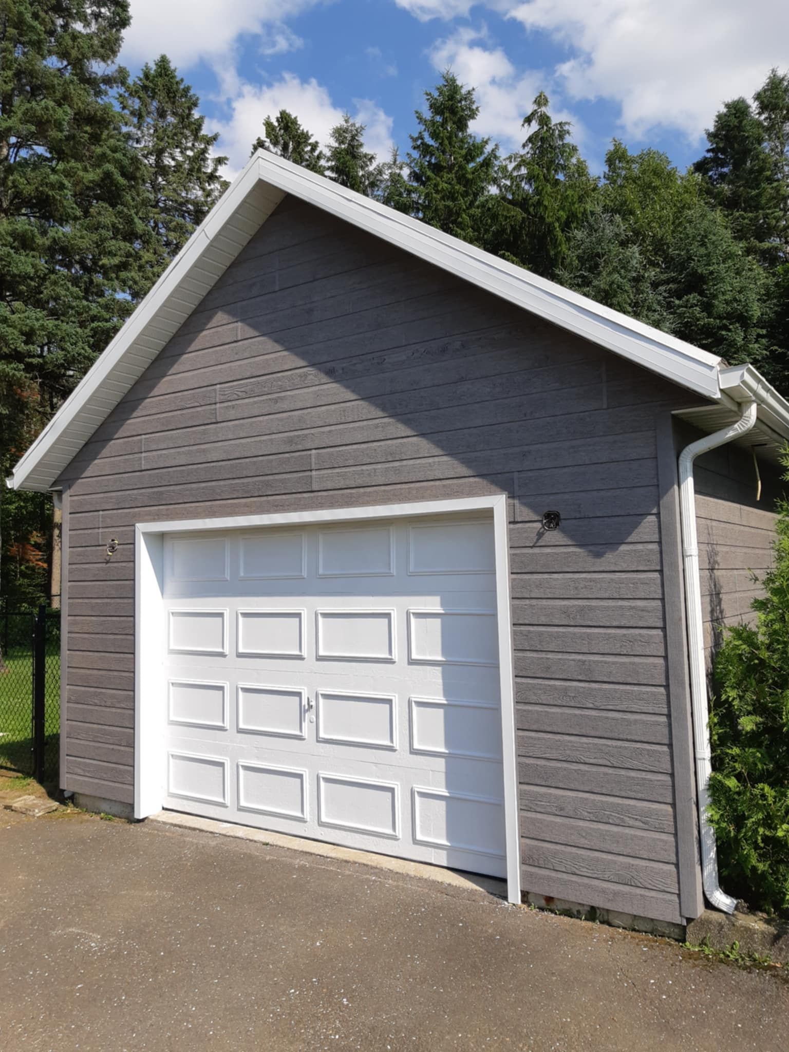 Garage gris avec porte et encadrements blancs, se détachant sur un fond d'arbres verts et de ciel bleu.