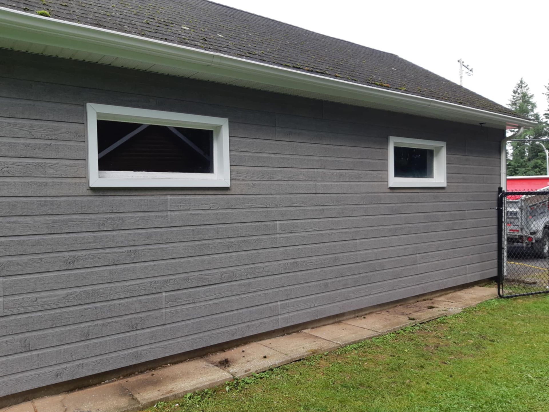 Bâtiment gris avec des fenêtres à encadrement blanc, un toit sombre et un carré d'herbe.