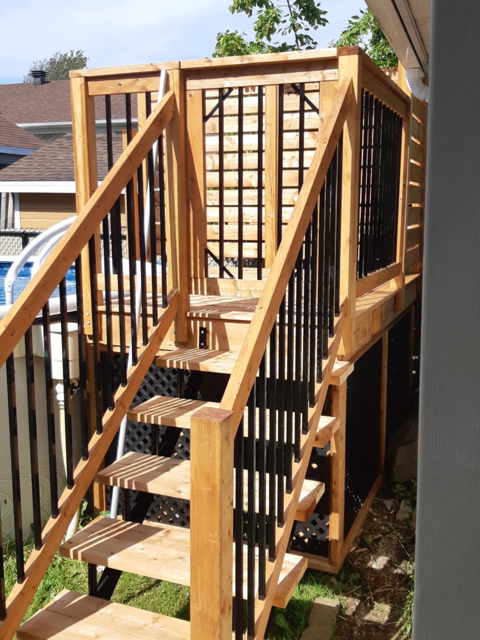 Terrasse en bois avec escalier, rambarde noire, fond en treillis, au bord d'une piscine.