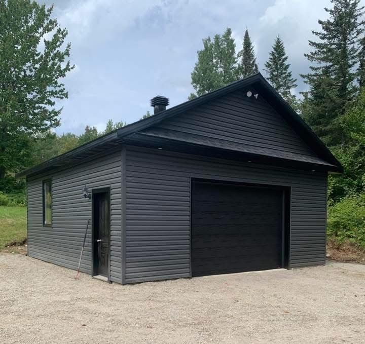 Garage gris avec porte noire, sur un sol gravillonné. Arbres en arrière-plan.