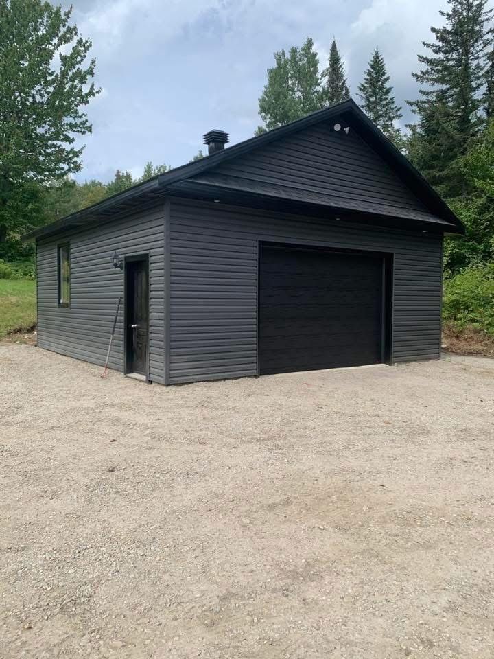 Bâtiment de garage gris avec porte de garage noire, situé sur une allée de gravier et entouré d'arbres.