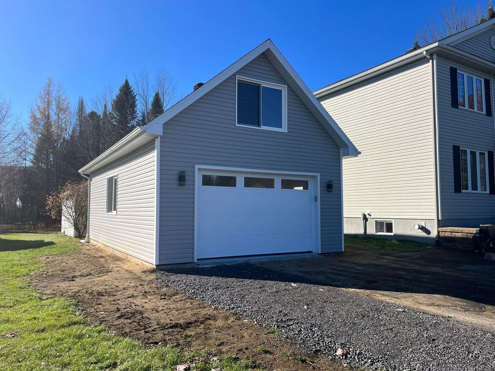 Garage indépendant aux murs gris, avec porte et fenêtre blanches. Allée gravillonnée devant, maison à côté.