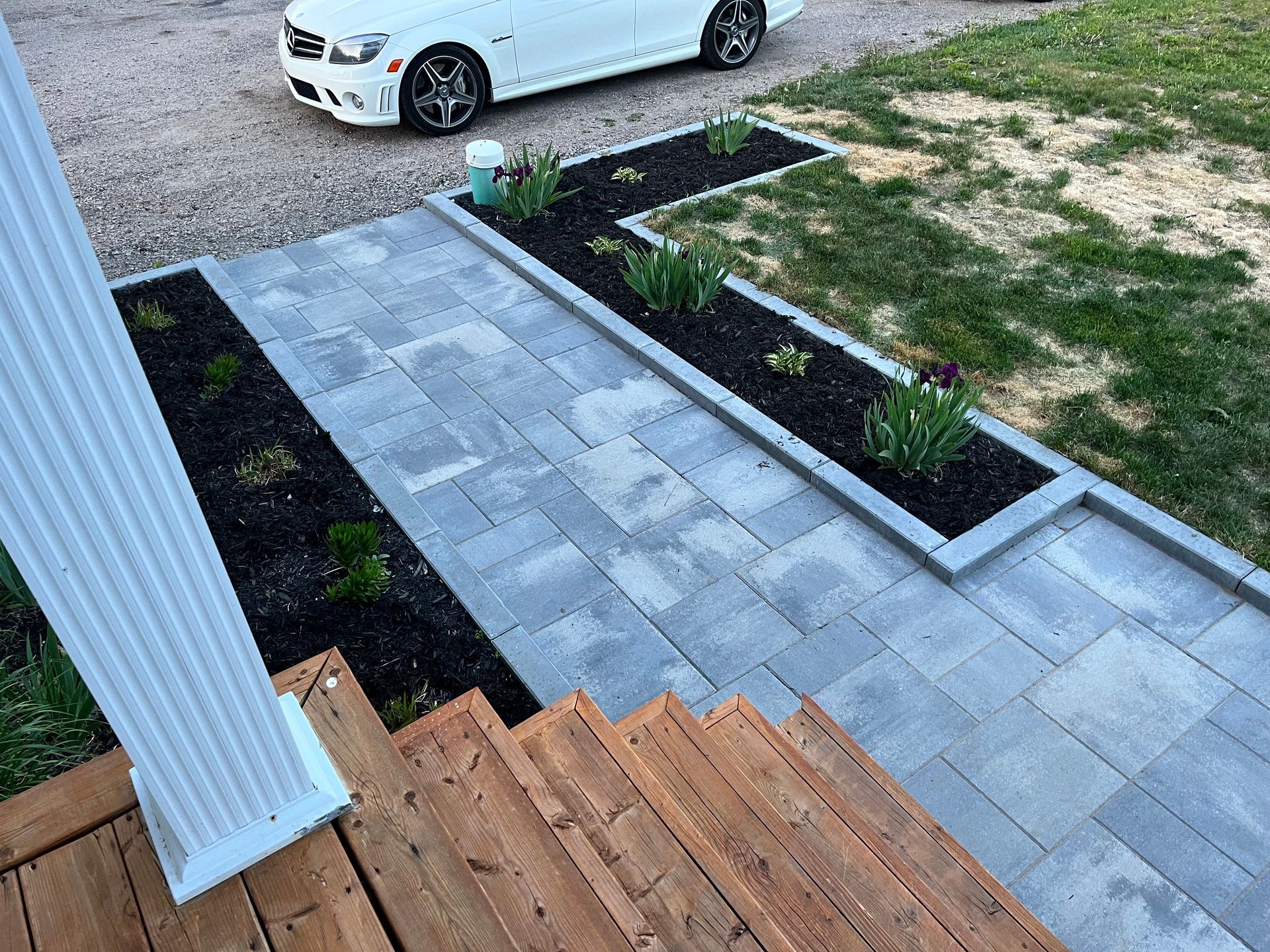 Allée de pierres et parterres de fleurs menant à des marches en bois, avec une voiture blanche visible en arrière-plan.
