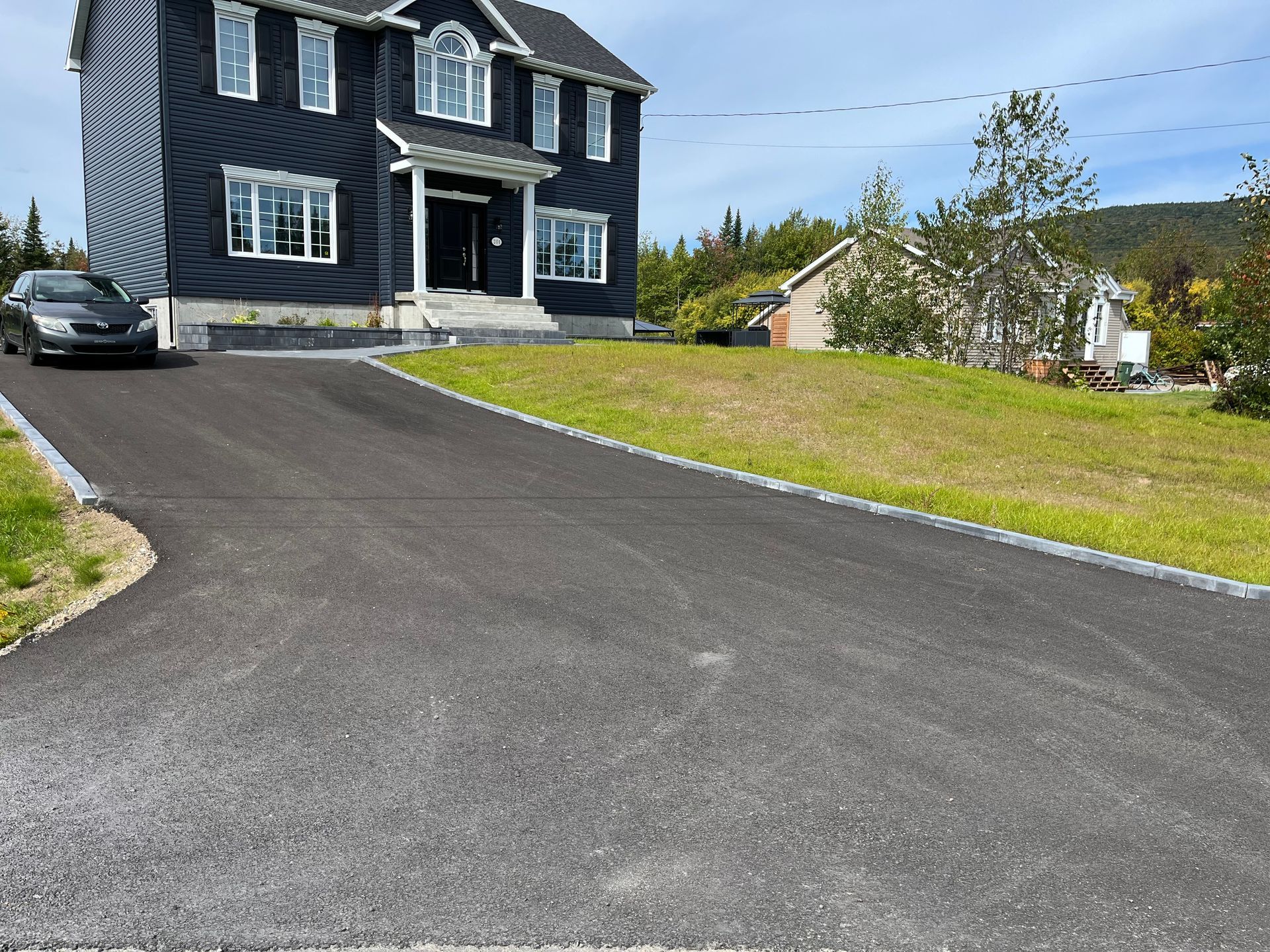 Maison noire avec allée pavée noire, voiture garée. Herbe verte et arbres en arrière-plan.