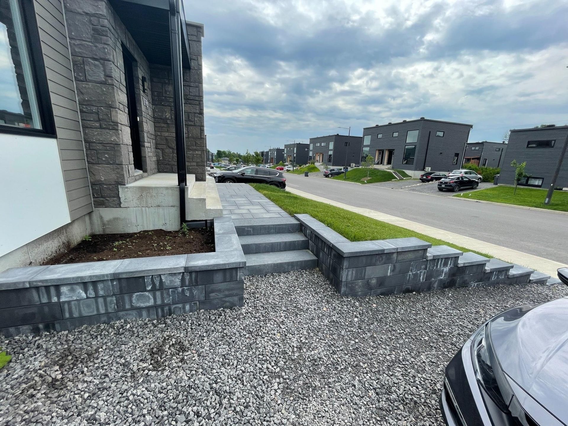 Entrée d'une maison en pierre grise avec des marches et un chemin de gravier, menant à une rue bordée de maisons similaires.