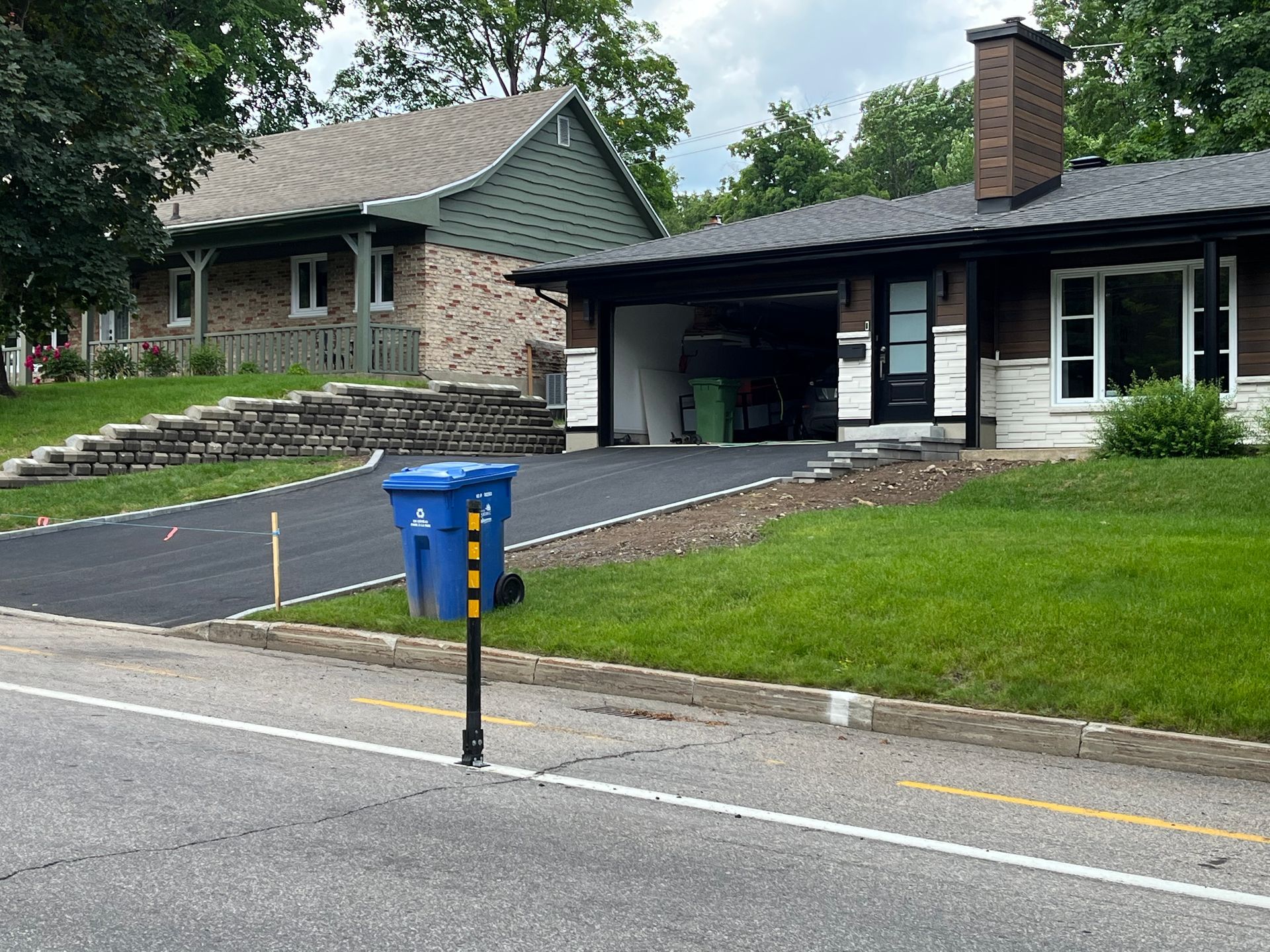 Deux maisons avec une allée nouvellement pavée ; une poubelle de recyclage bleue sur un poteau en bord de route.