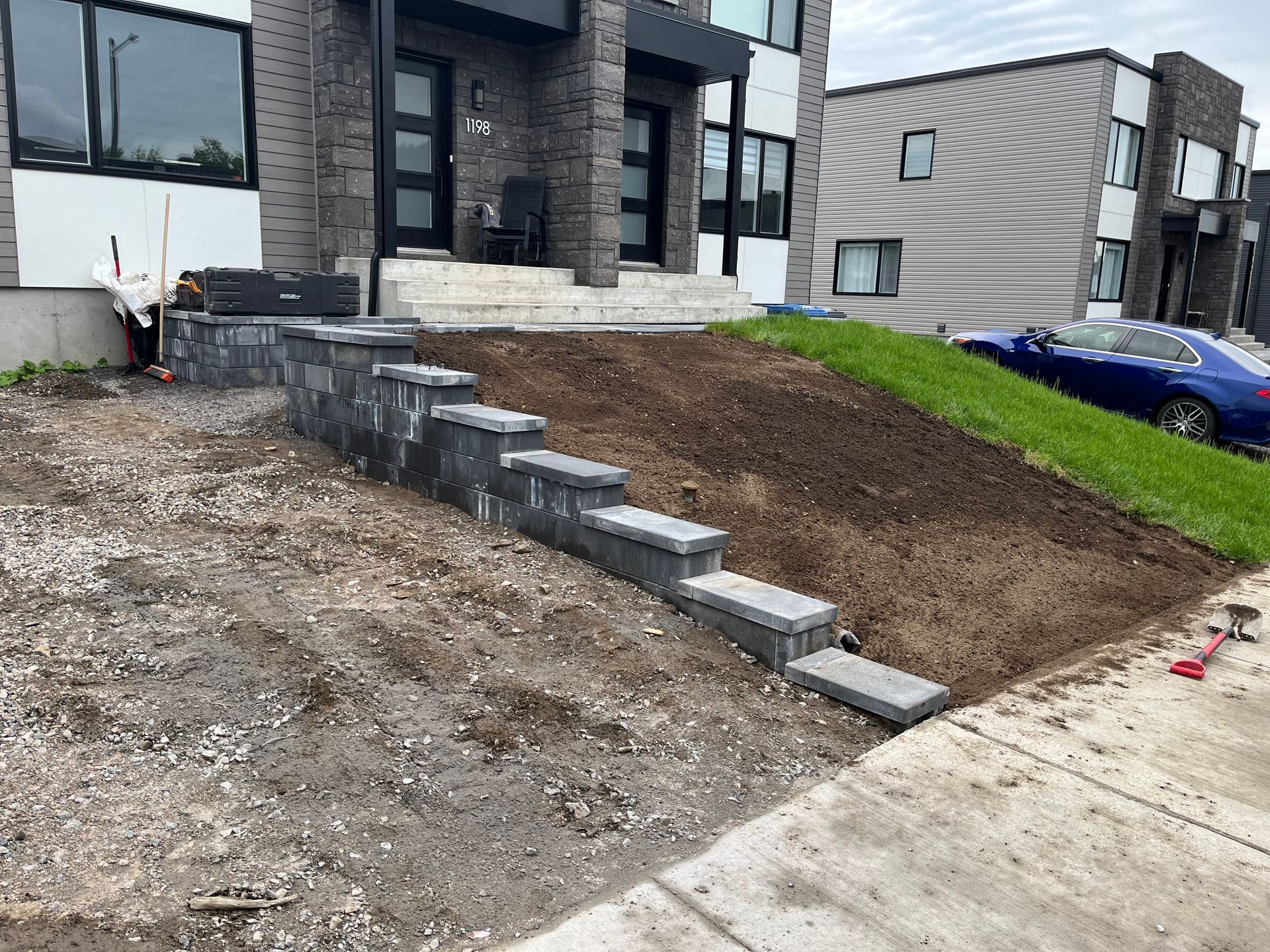 Des marches en briques mènent à une maison moderne avec une porte noire. Un jardin en pente, bordant les marches, est en cours d'aménagement paysager.
