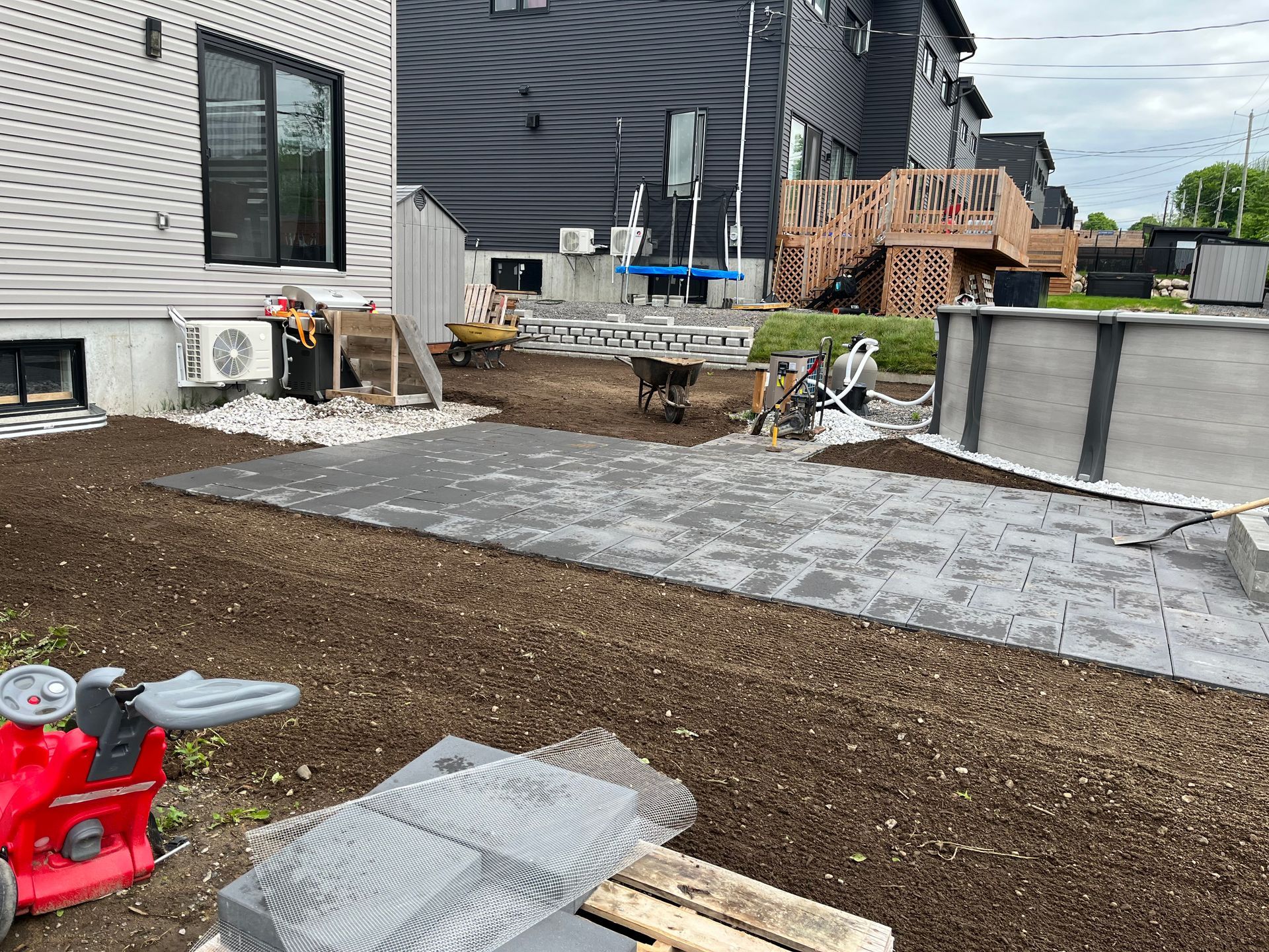 Jardin arrière en travaux : dalles grises posées, gravier, outils, maison en arrière-plan.