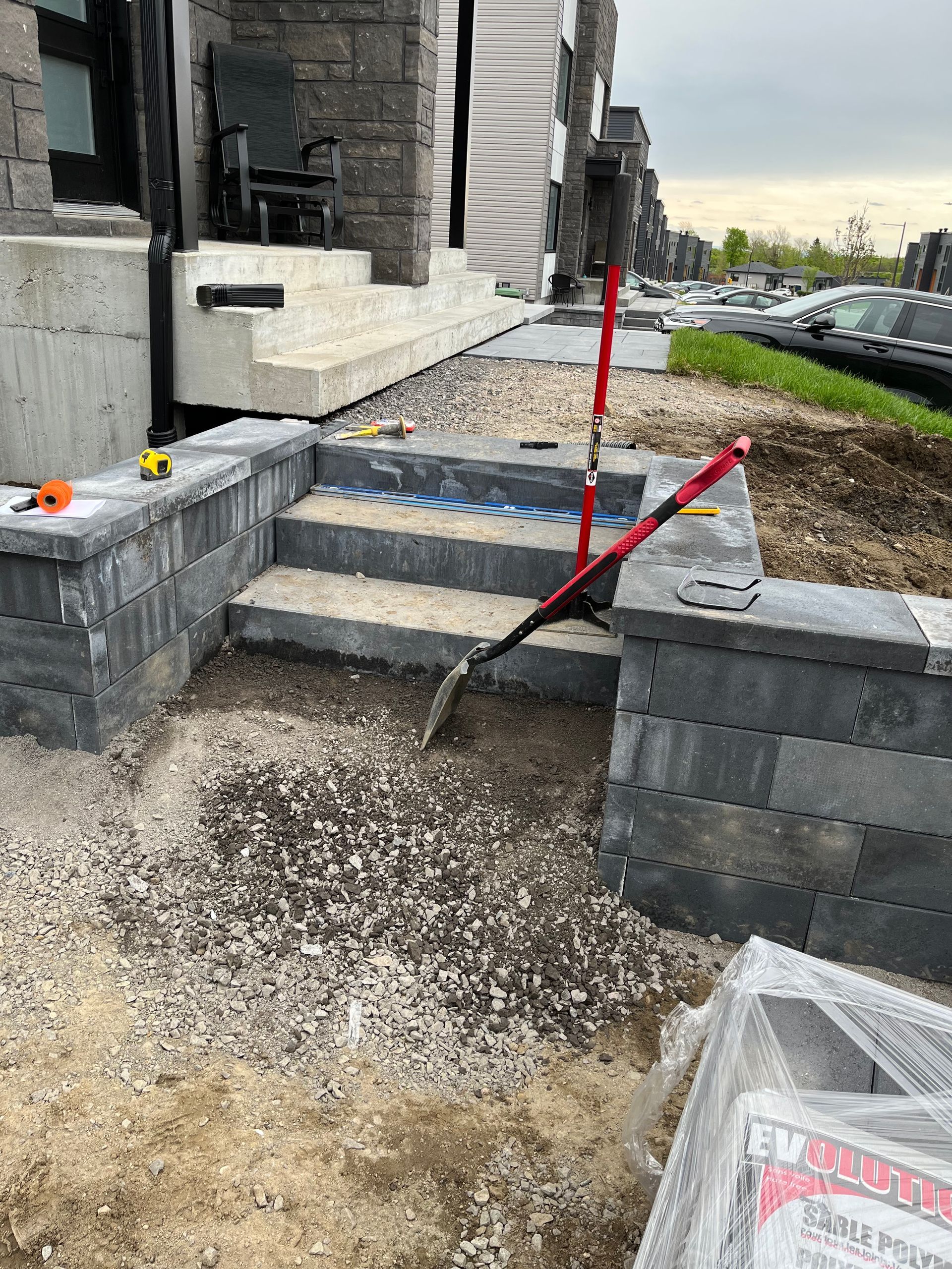 Des marches en pierre sont en construction à l'entrée d'un bâtiment. Un outil rouge, du gravier et des blocs sont visibles.