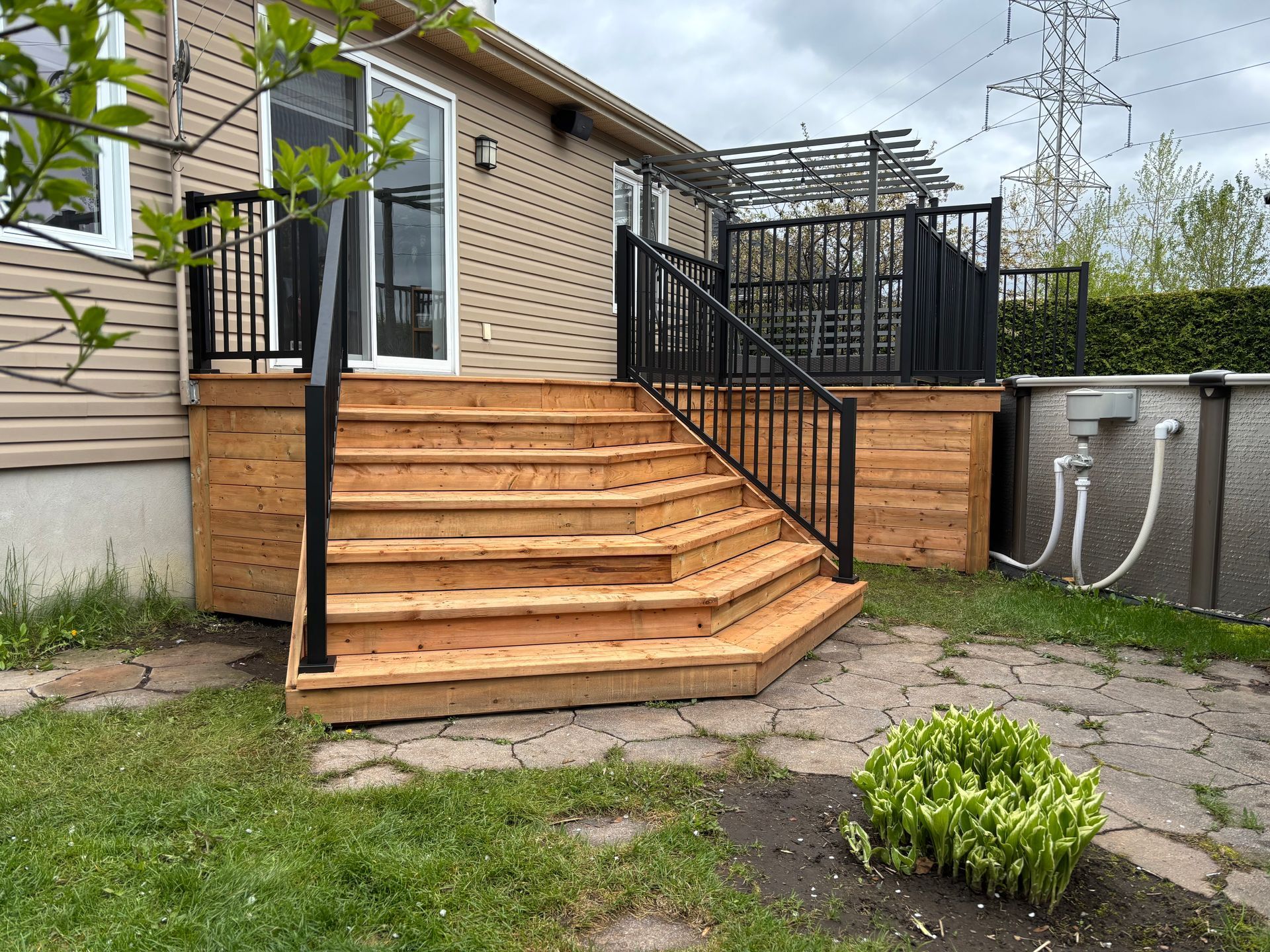 Terrasse en bois avec escalier, rambarde noire et porte-fenêtre coulissante. Une piscine est visible sur la droite.