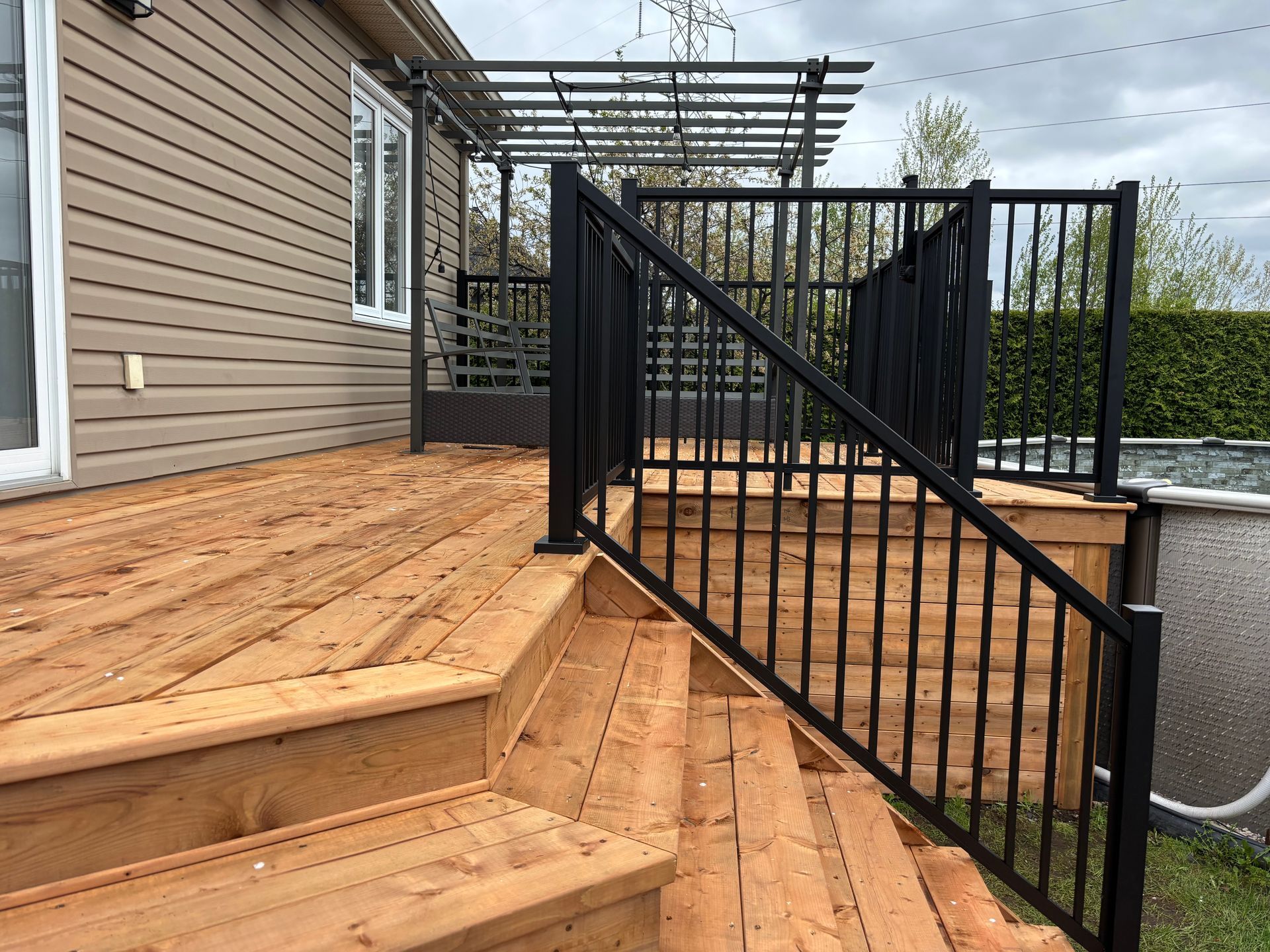 Terrasse en bois avec rambarde noire et marches menant à un coin salon avec une pergola.