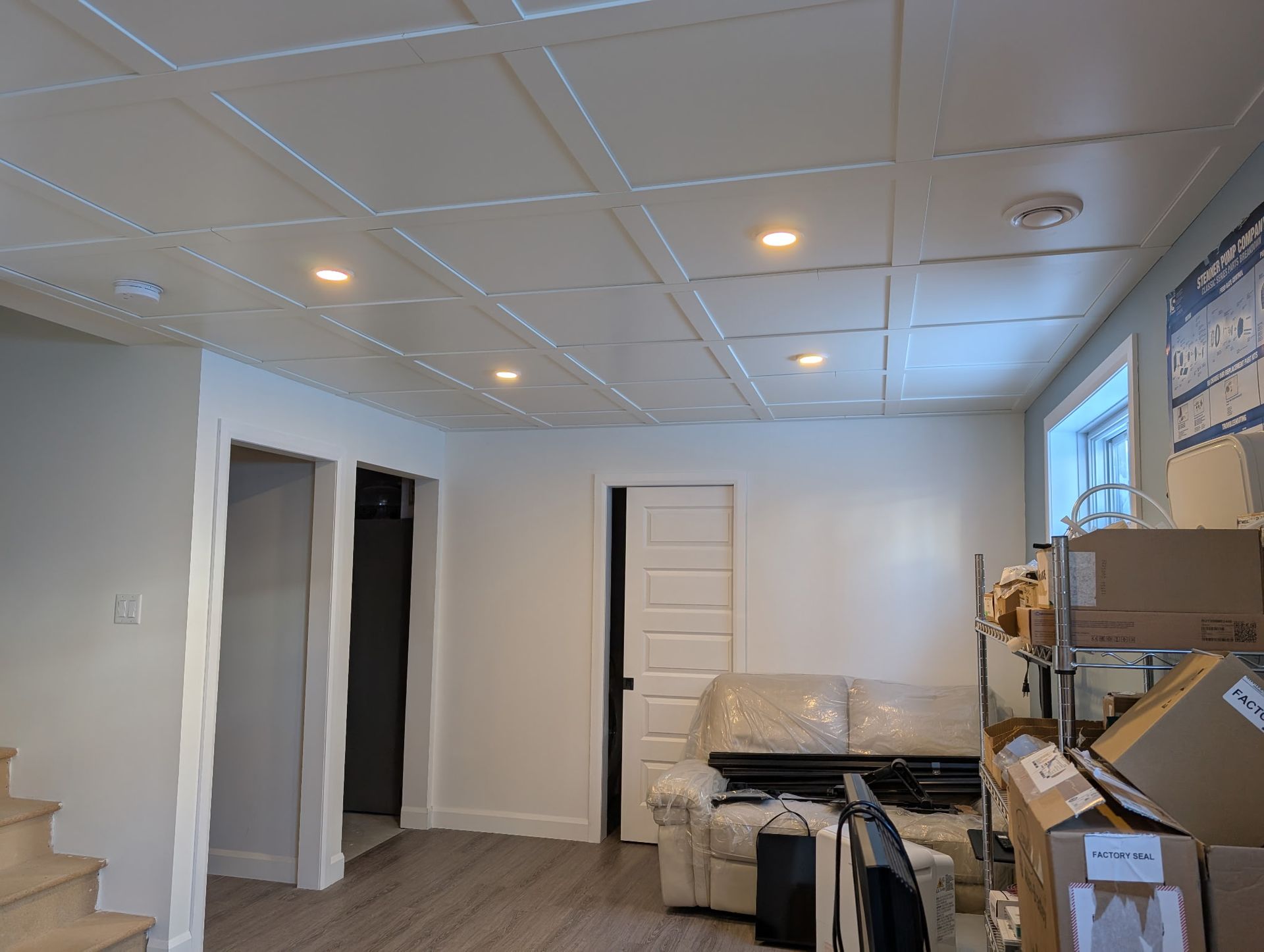 Pièce aux murs blancs avec faux plafond, spots encastrés, porte ouverte et meubles adossés à un mur avec une fenêtre.