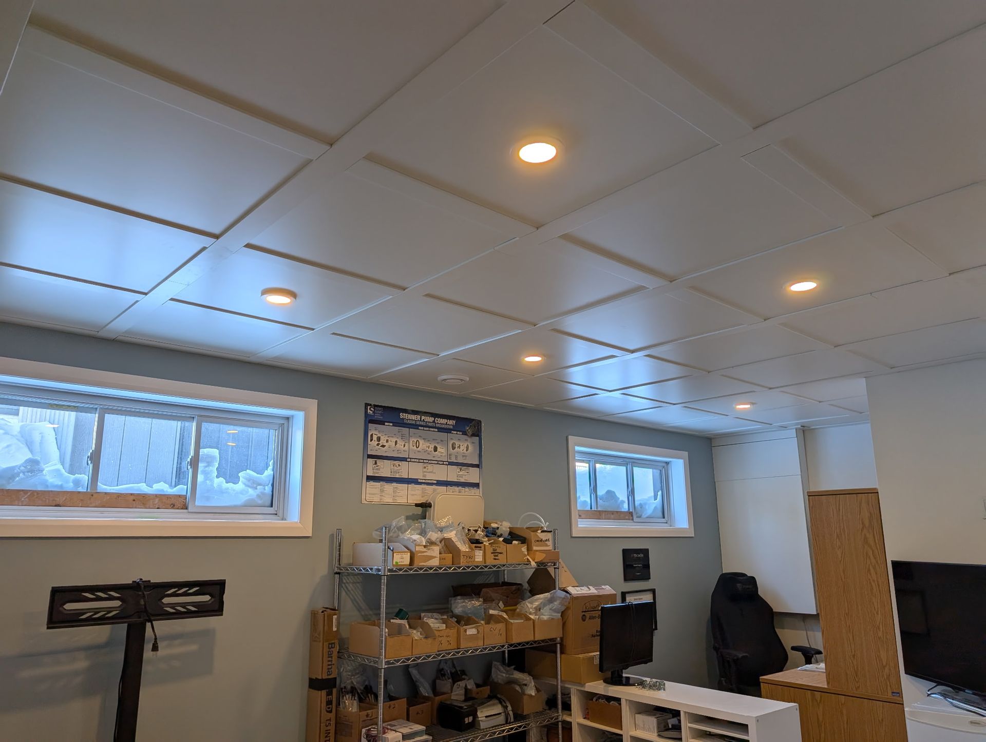 Plafond à panneaux blancs avec éclairage encastré, au-dessus d'un espace de travail en sous-sol avec fenêtres et étagères.