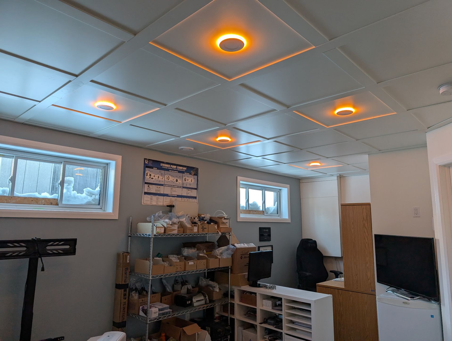 Pièce au sous-sol avec plafond en dalles bleues et spots encastrés orange ; fenêtres, étagères, bureau, boîtes.