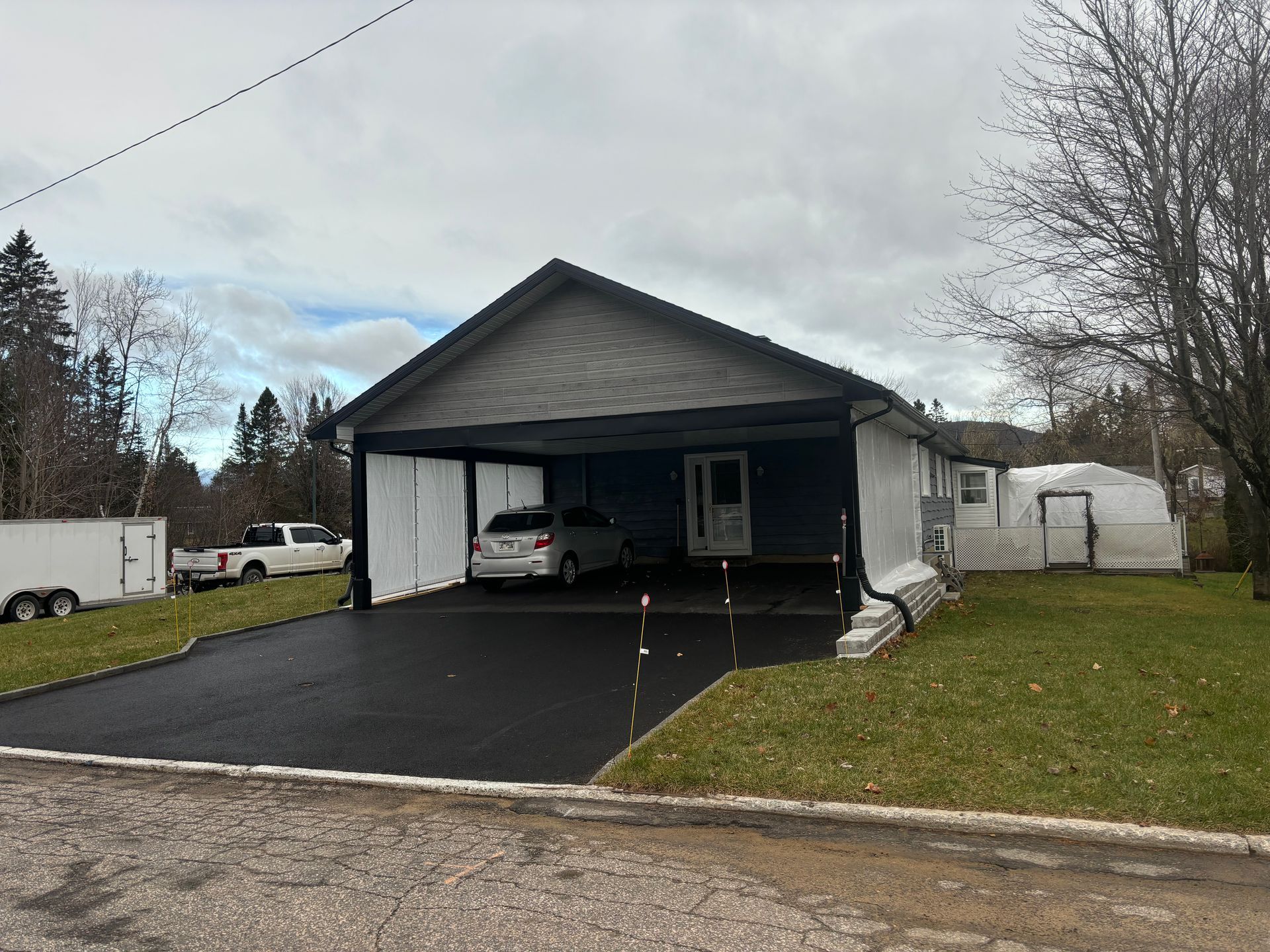 Abri voiture attenant à une maison bleue avec une voiture garée à l'intérieur. Allée en asphalte noir, ciel couvert.