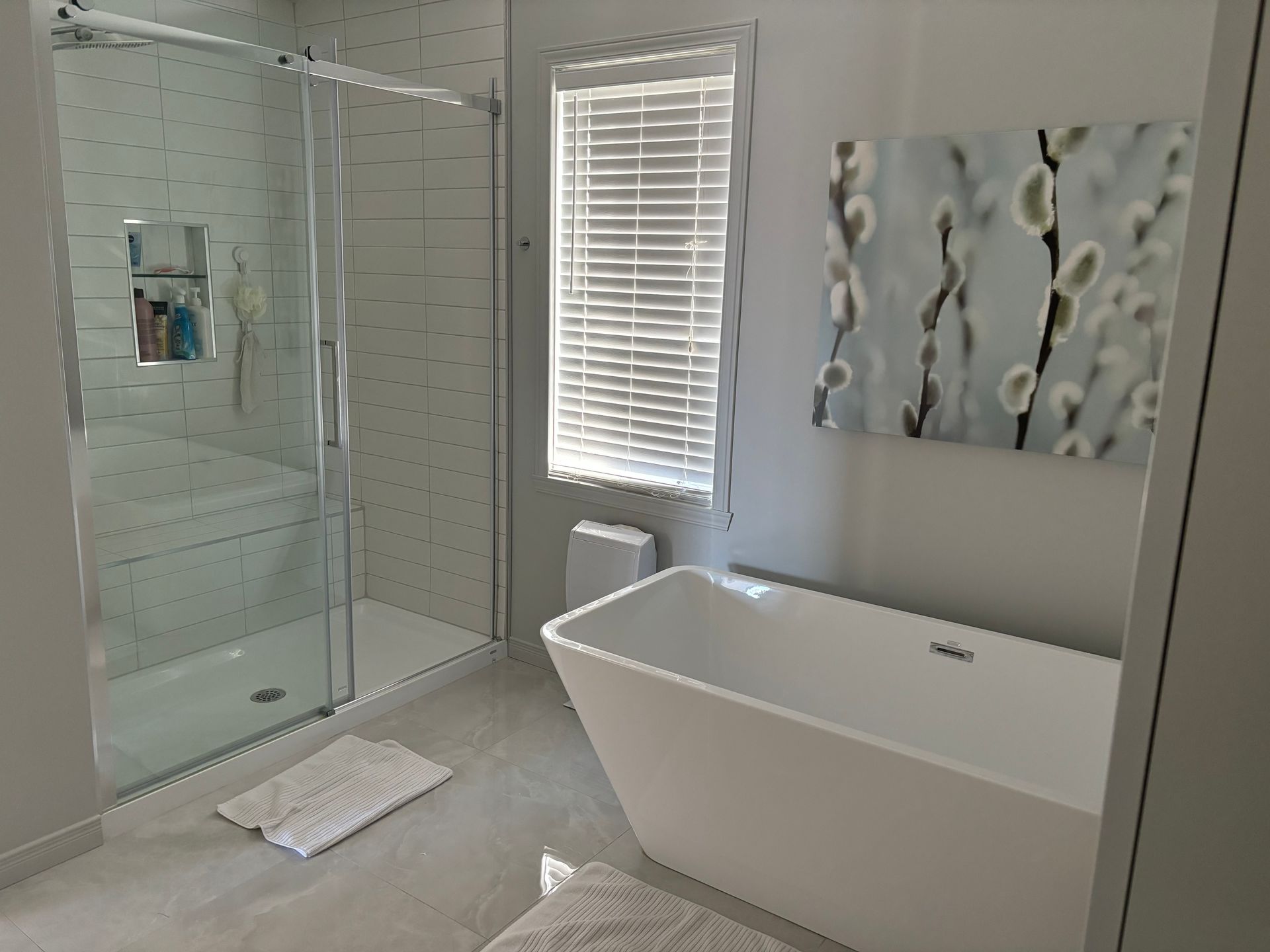 Salle de bains moderne avec douche en verre, baignoire et fenêtre à stores. Couleurs douces et décoration minimaliste.