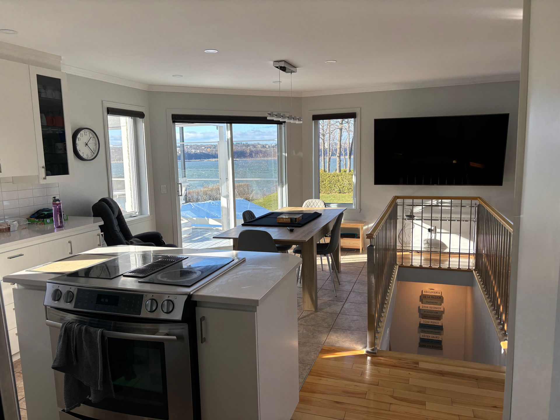 Cuisine avec vue sur l'eau, ouverte sur la salle à manger et l'escalier. Électroménagers en acier inoxydable, grande télévision et table.