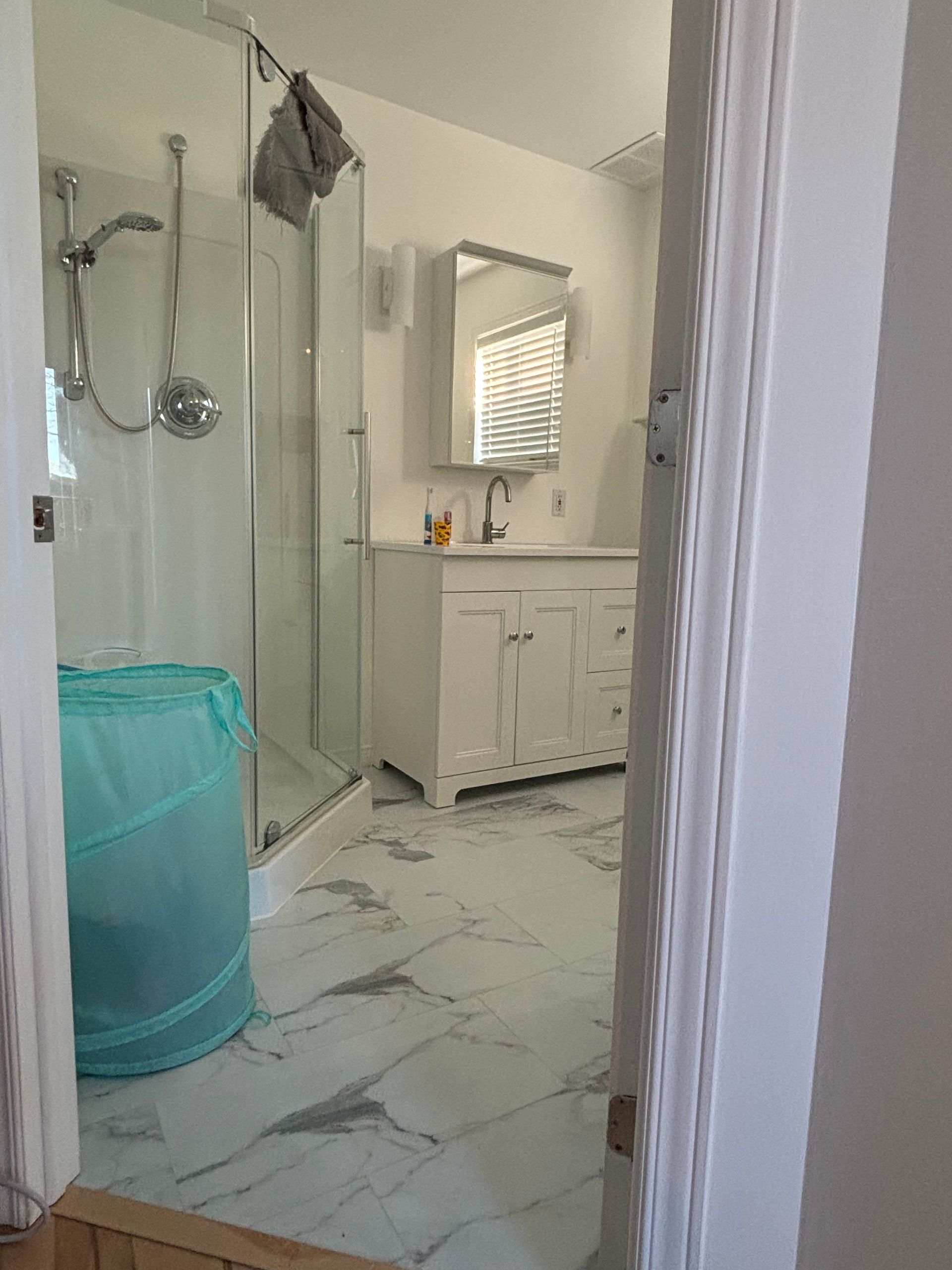 Intérieur de salle de bains avec meuble-lavabo blanc, sol en marbre et douche. Un panier à linge est posé à gauche.