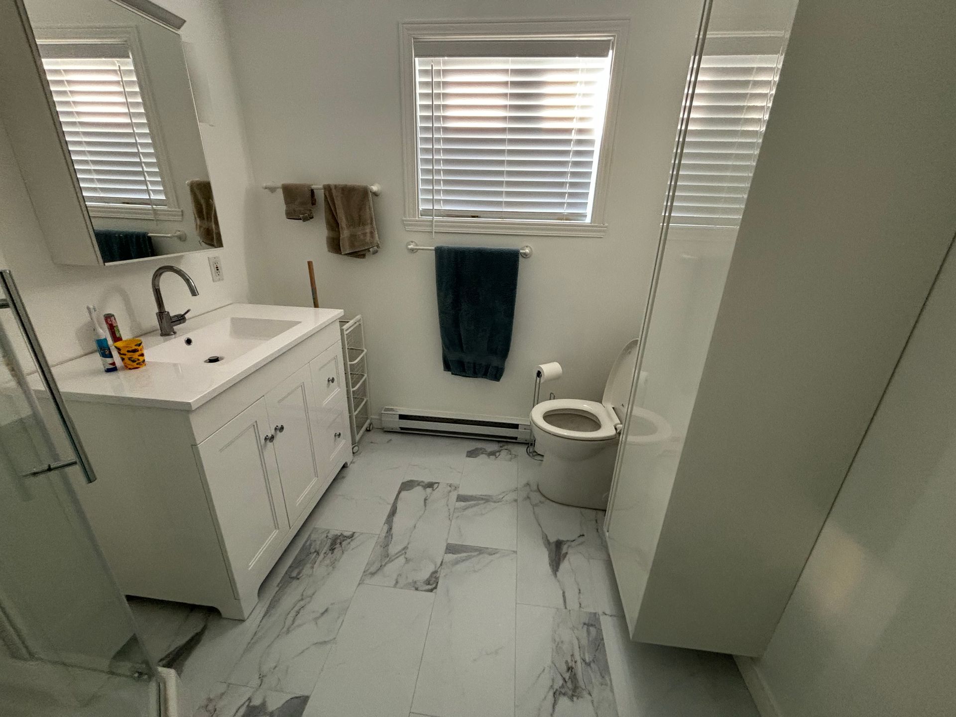 Salle de bains avec meuble-lavabo, toilettes et sol blancs ; porte-serviettes bleu, stores à la fenêtre.