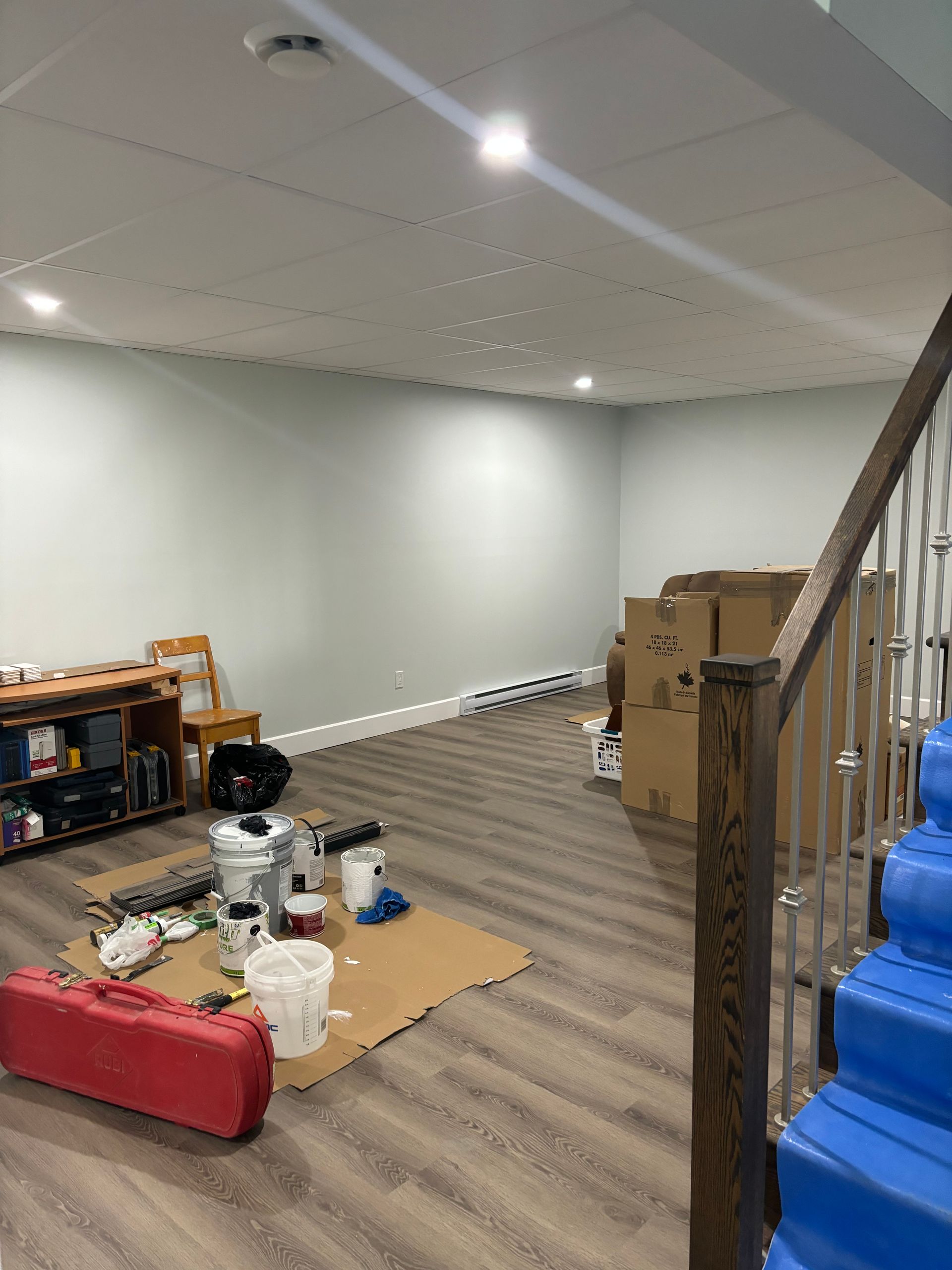 Pièce au sous-sol en cours de rénovation avec matériel de peinture, cartons et escalier. Murs et sol clairs.
