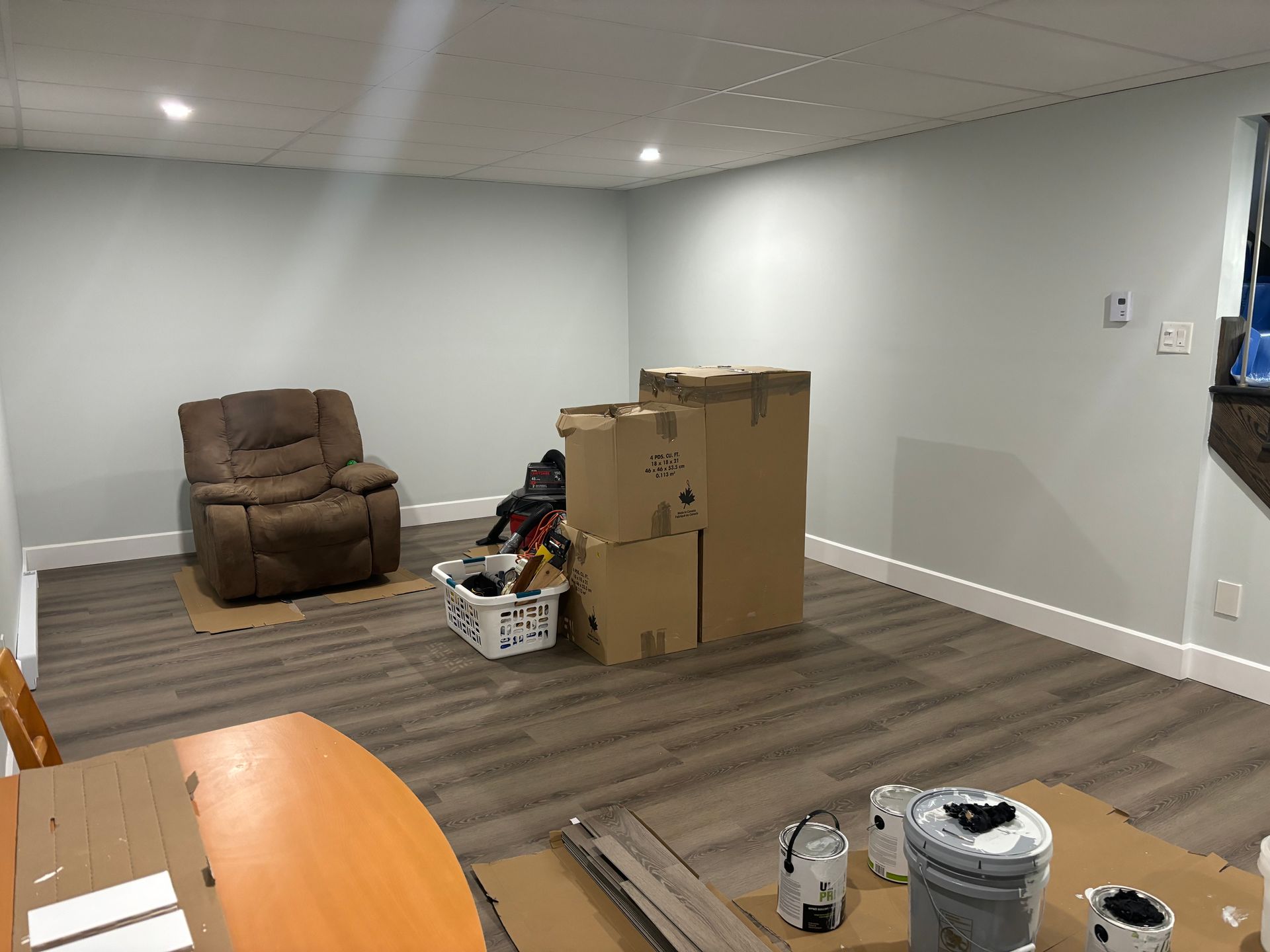 Une pièce presque vide, avec des murs gris, un sol imitation bois, un fauteuil inclinable et des cartons empilés.