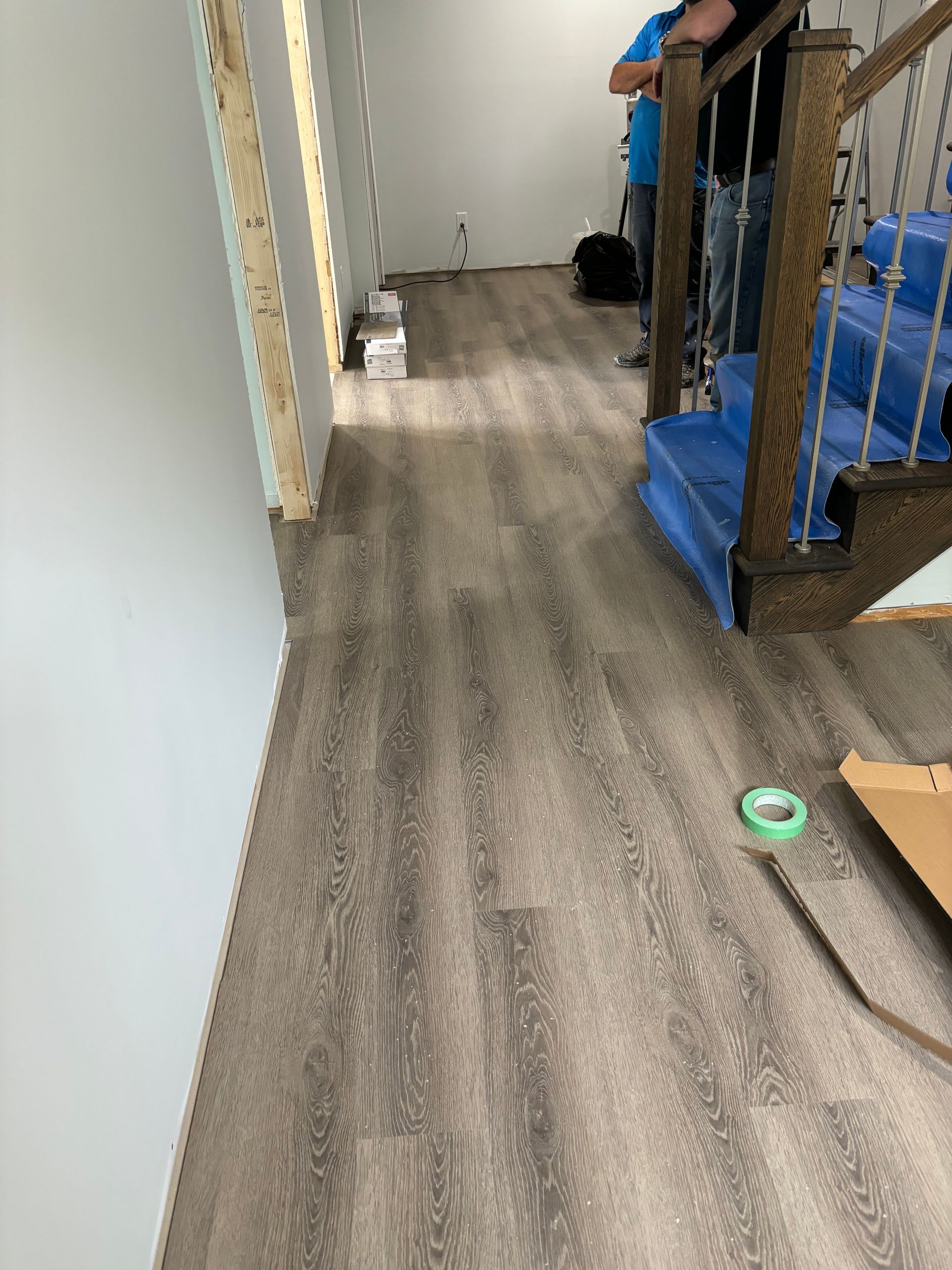 Revêtement de sol imitation bois récemment installé dans un couloir avec escalier et porte. Des personnes se tiennent à proximité.