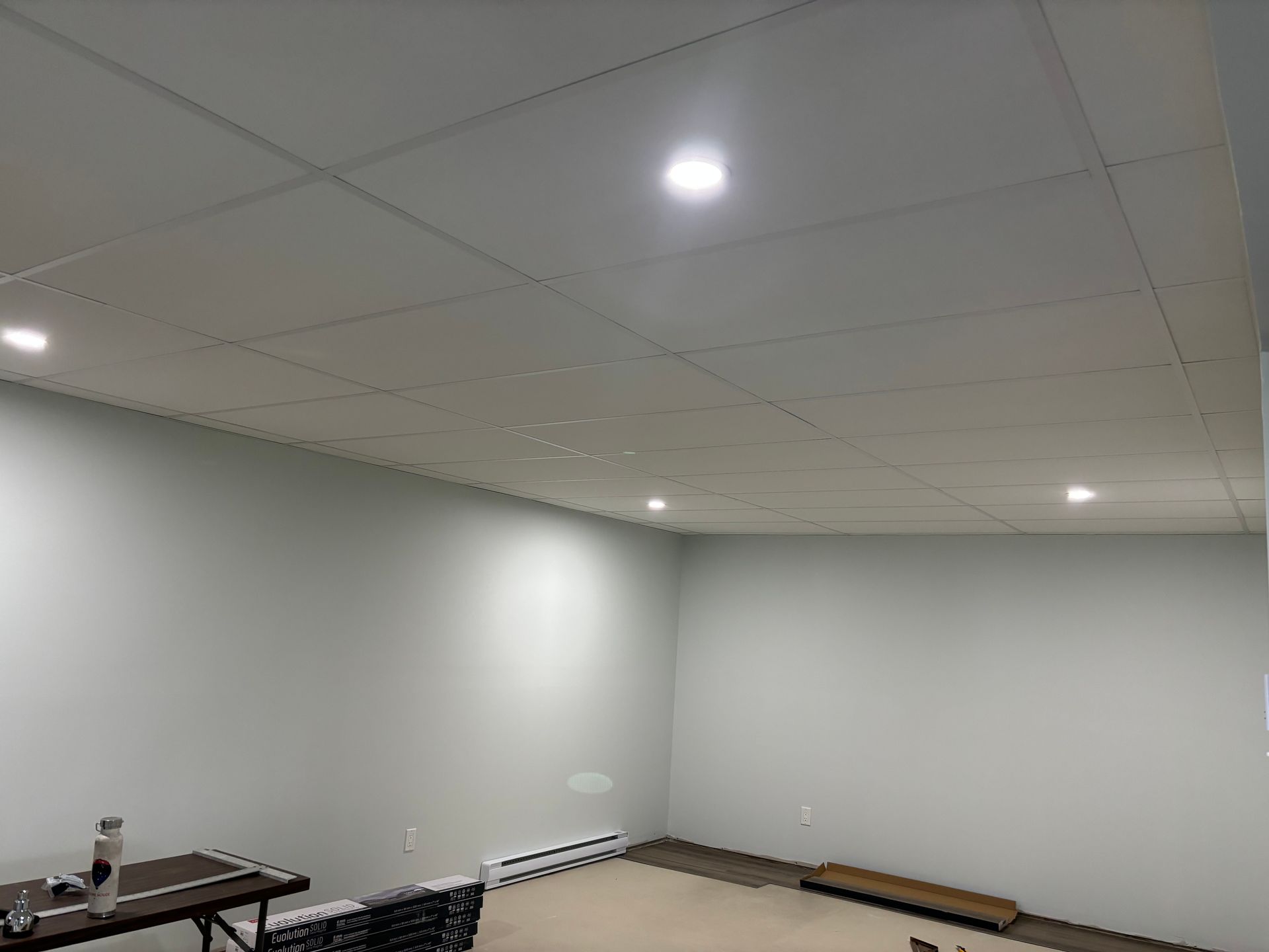 Une pièce aux murs blancs, un plafond à grille avec des spots encastrés et une table avec une bouteille d'eau.