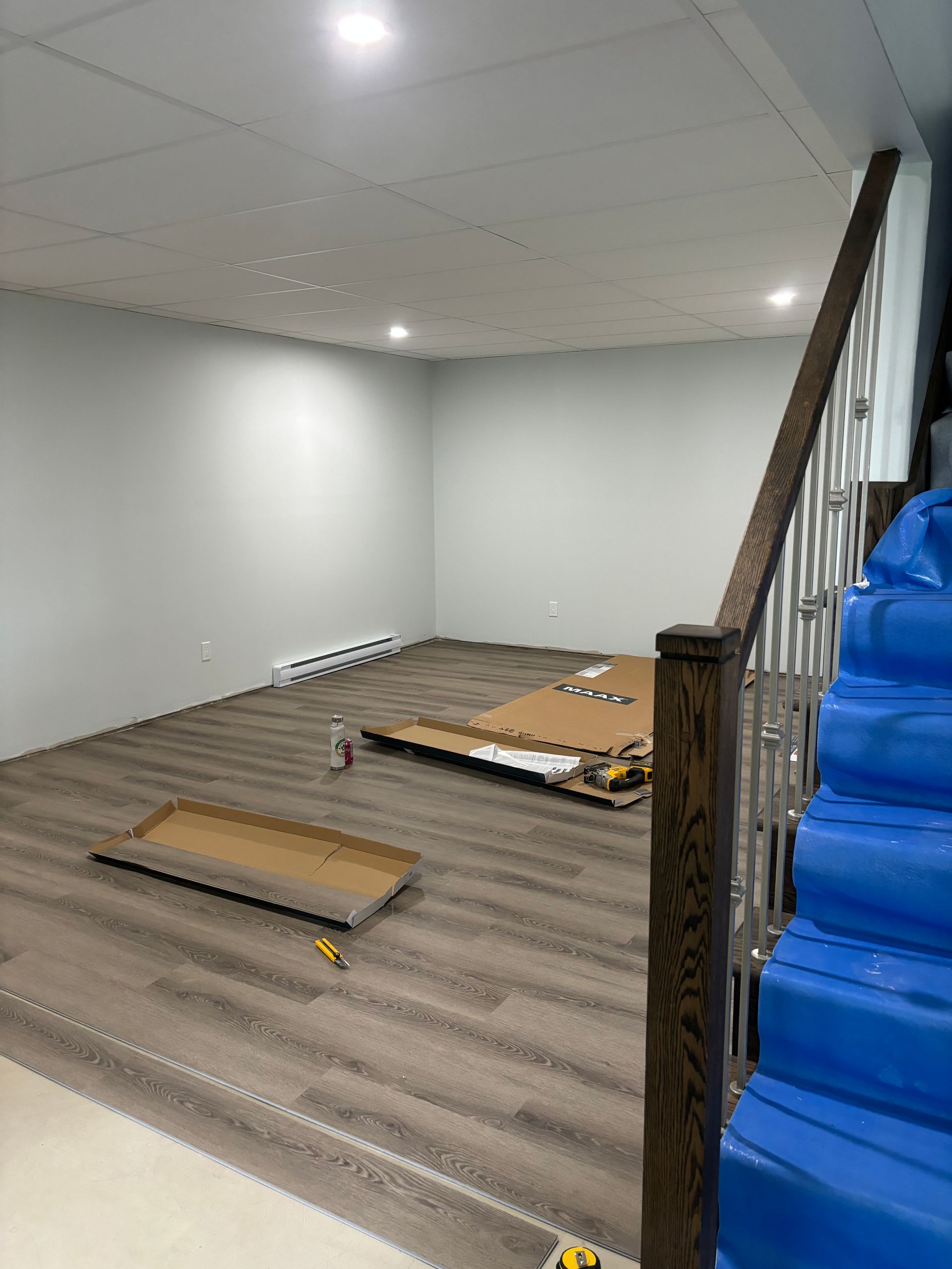 Pièce vide avec sol gris, murs blancs et rampe en bois. L'escalier à droite a des marches recouvertes de tissu bleu.