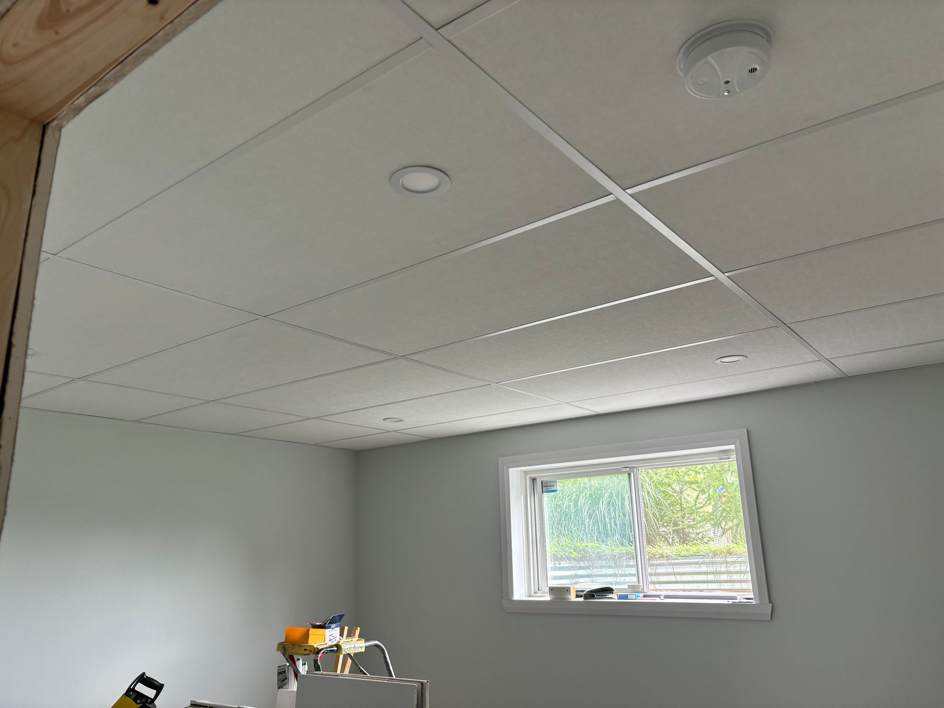 Plafond carrelé blanc dans une pièce avec une fenêtre, des murs en plaques de plâtre et quelques outils.