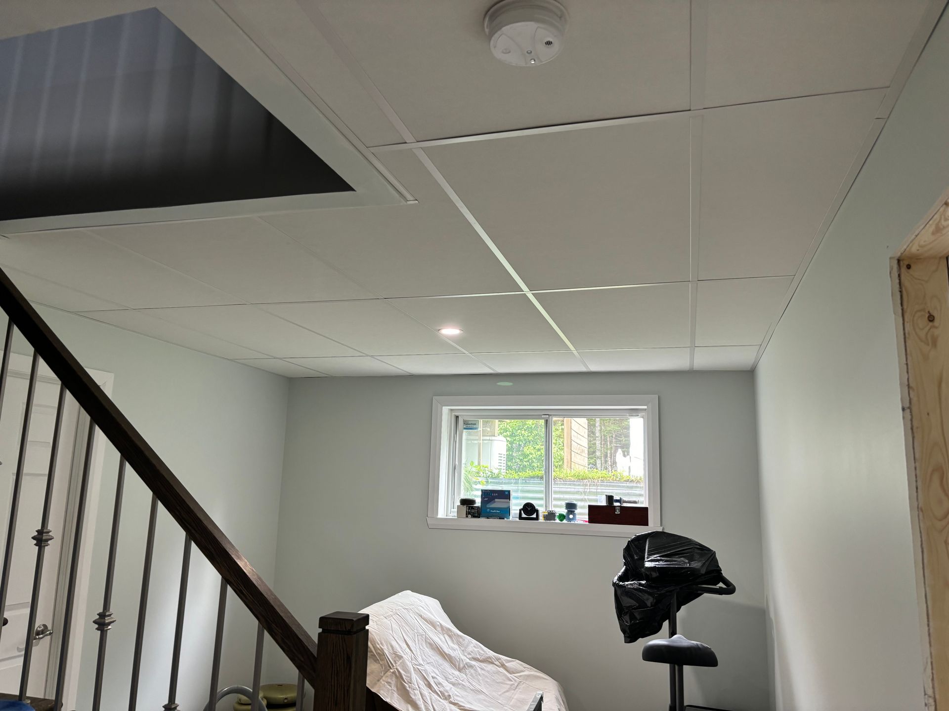 Intérieur du sous-sol avec faux plafond, escalier, fenêtre et détecteur de fumée. Dominante de couleurs blanc et bleu clair.