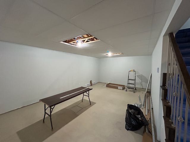 Sous-sol non aménagé avec trous au plafond. Table et échelle présentes. Murs et sol clairs. Escalier à droite.