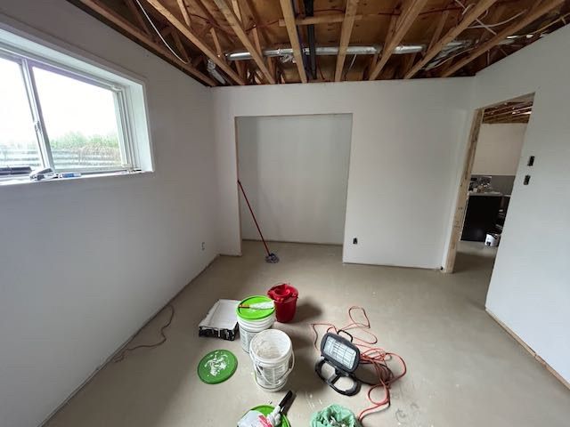 Pièce intérieure en travaux, avec des murs blancs peints, une fenêtre et des poutres apparentes au plafond. Matériel de peinture au sol.