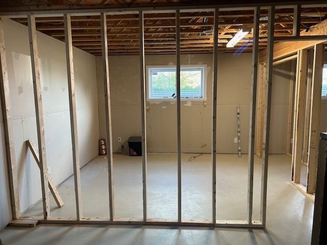 Ossature intérieure d'une pièce avec fenêtre, sol en béton et solives de plafond apparentes. Les murs sont partiellement revêtus.