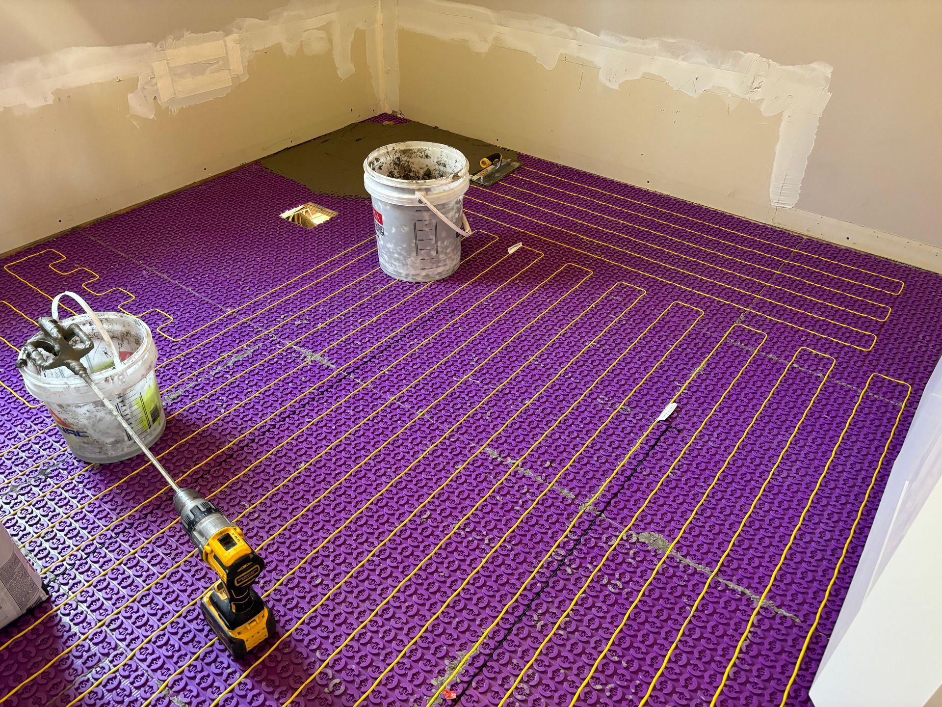 Un tapis chauffant violet pour le sol est posé dans une pièce, avec un seau et des outils à proximité.