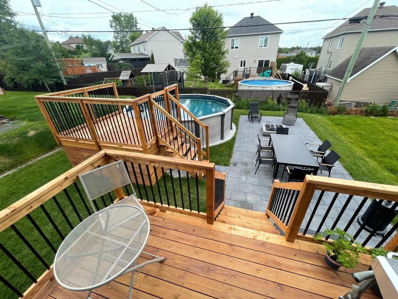 Cour arrière avec terrasse en bois, piscine hors sol, mobilier de jardin et pelouse verdoyante. Quartier résidentiel en arrière-plan.