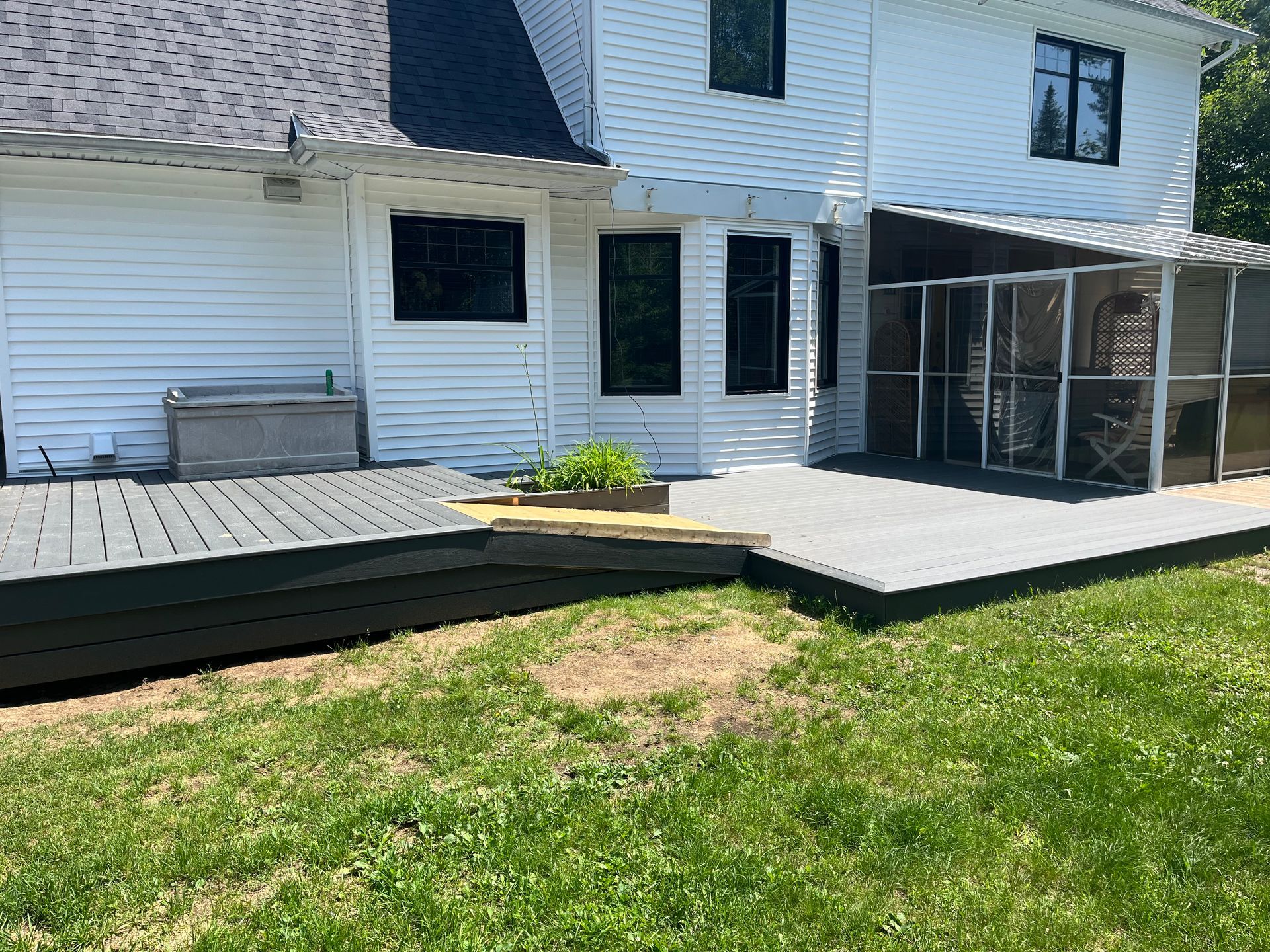 Terrasse arrière avec des planches gris foncé, attenante à une maison blanche, et véranda grillagée.